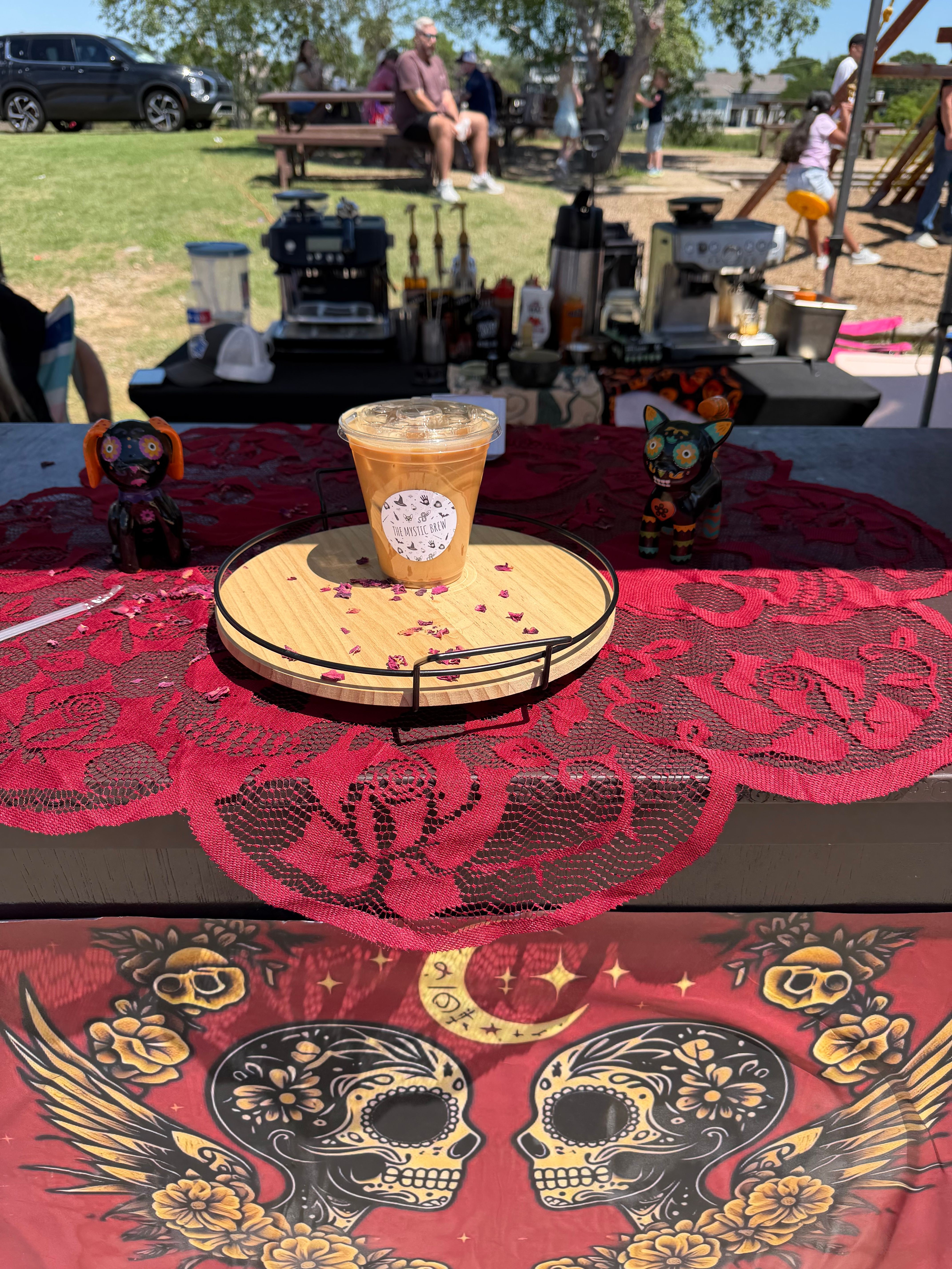 A table with a drink and a cake, decorated with a red cloth, outdoors with people and equipment in the background.