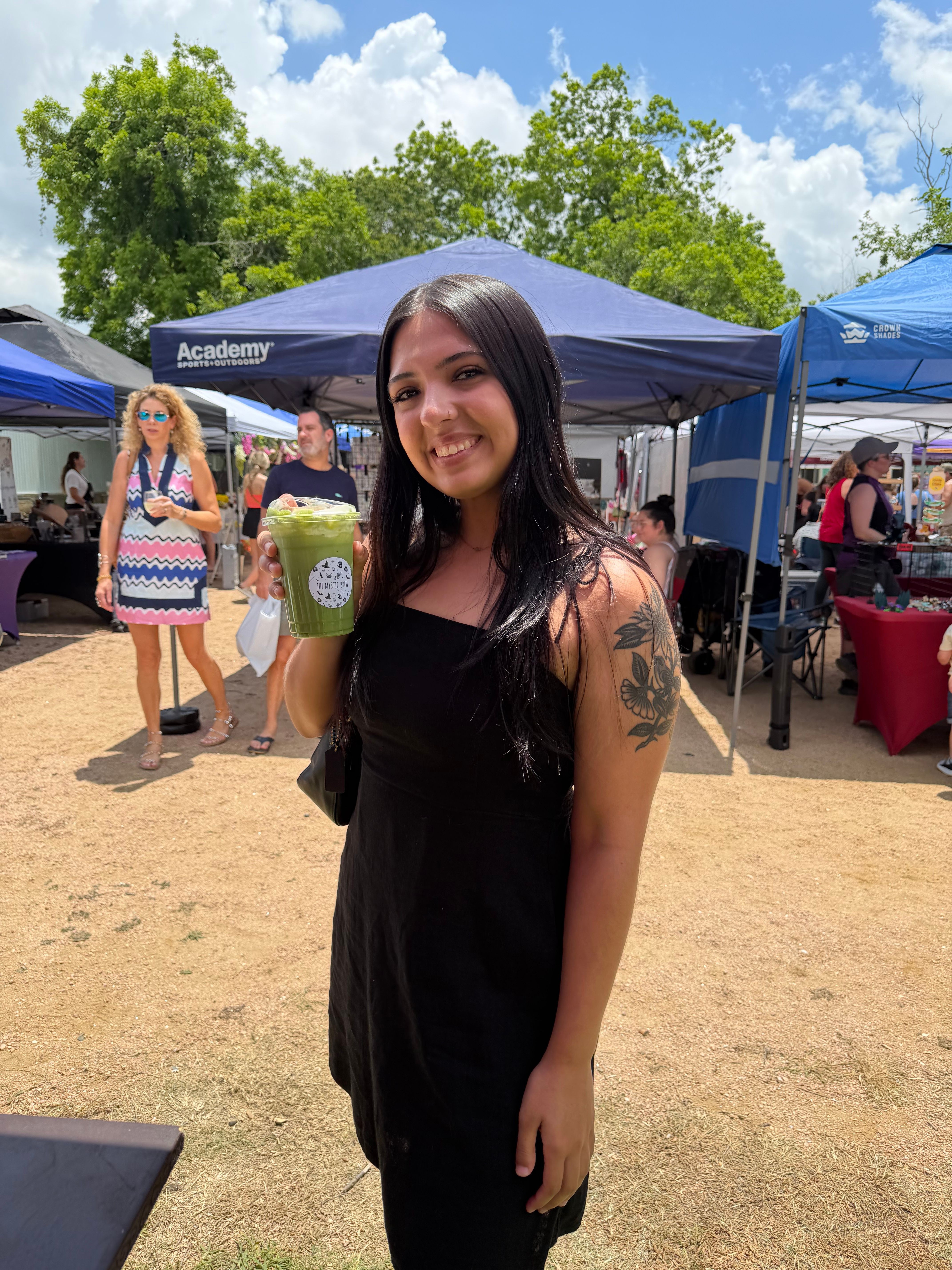 Woman with long dark hair in a black dress holding a drink at an outdoor event, with tents and people in the background.