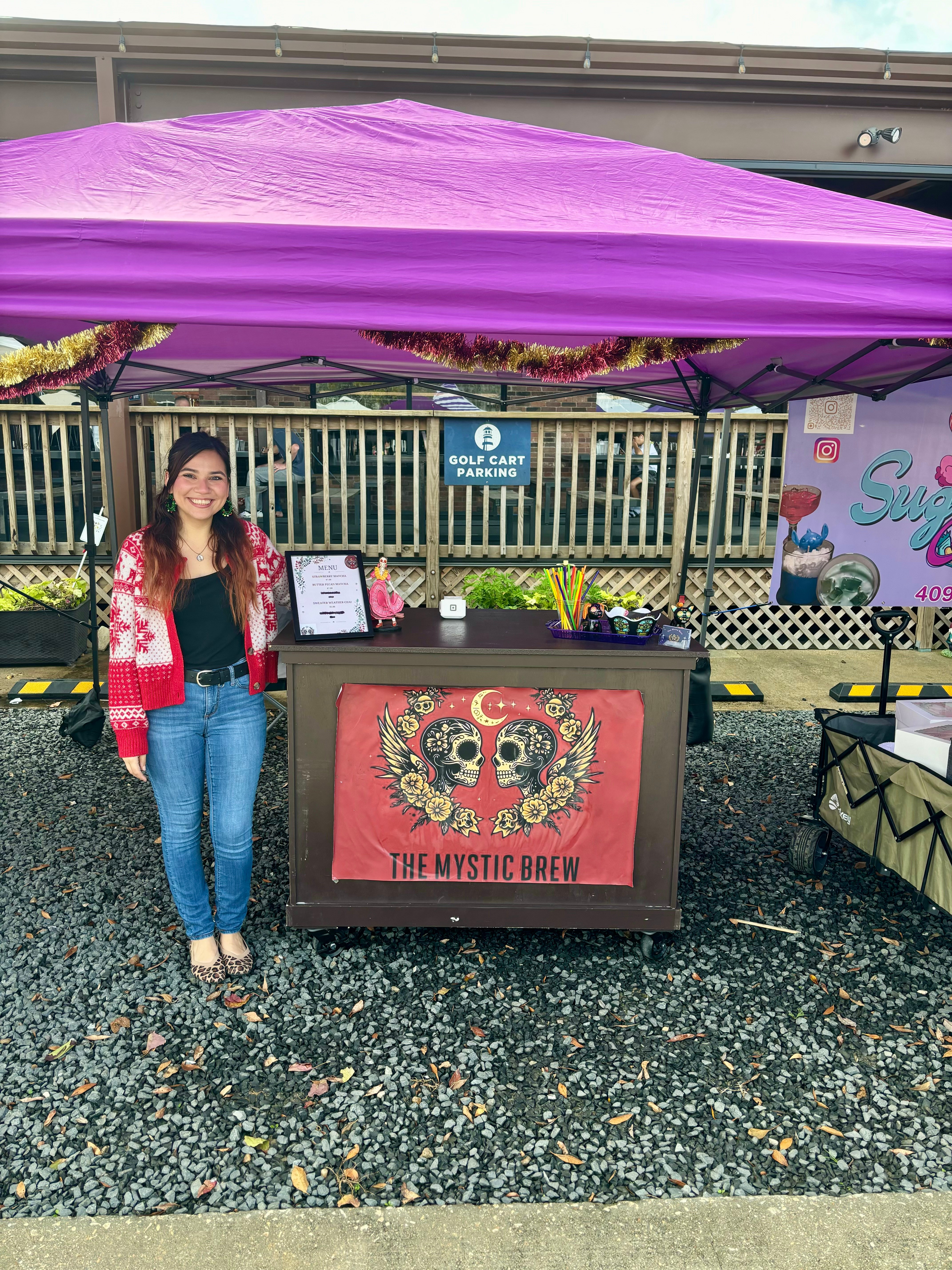 Woman standing next to a booth with a purple canopy, decorated with a colorful sugar skull banner, outdoors on a rainy day.