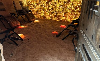 Indoor space with chairs, illuminated salt wall, sandy floor, and a partially open door on the right.