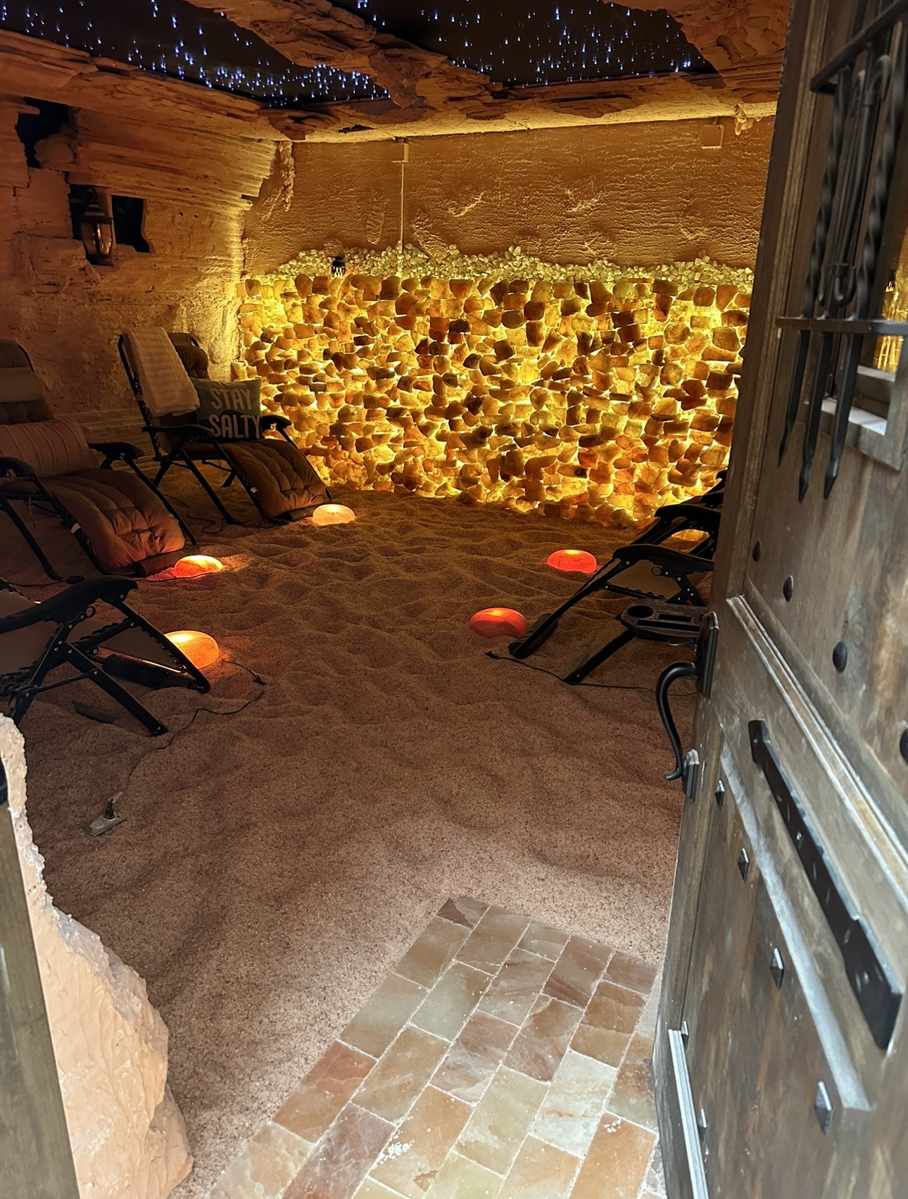 Indoor space with chairs, illuminated salt wall, sandy floor, and a partially open door on the right.