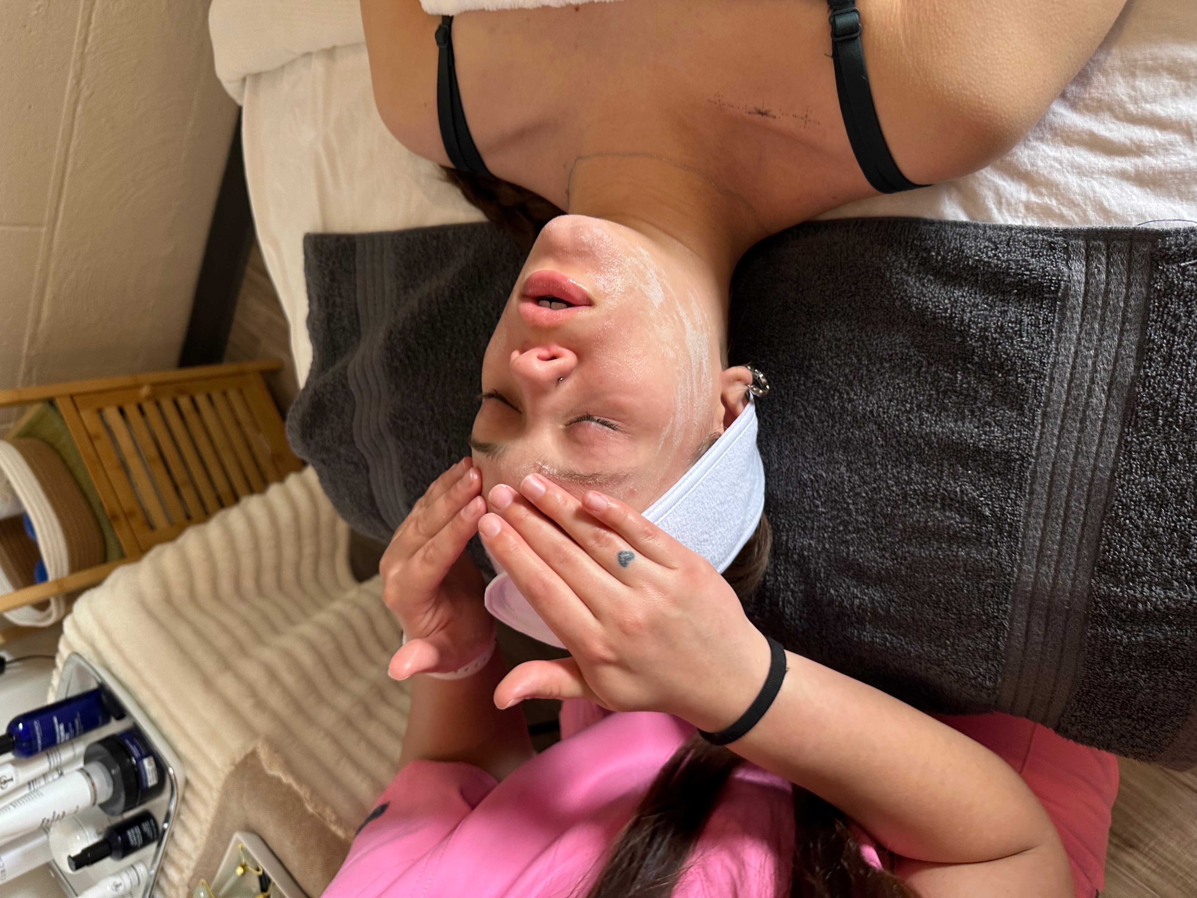 Person lying on bed with head tilted back, eyes closed, hands on face, wearing a headband and black straps.