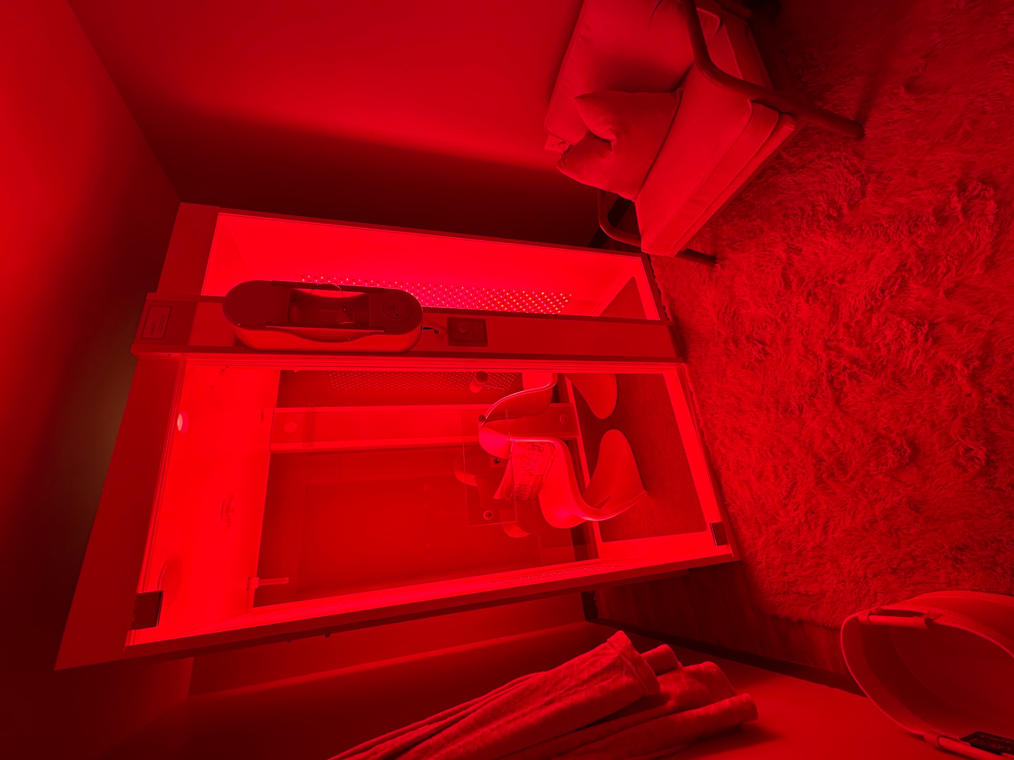 Room with red lighting, mirror, and a bed with a blanket, viewed from the side, with a carpeted floor.