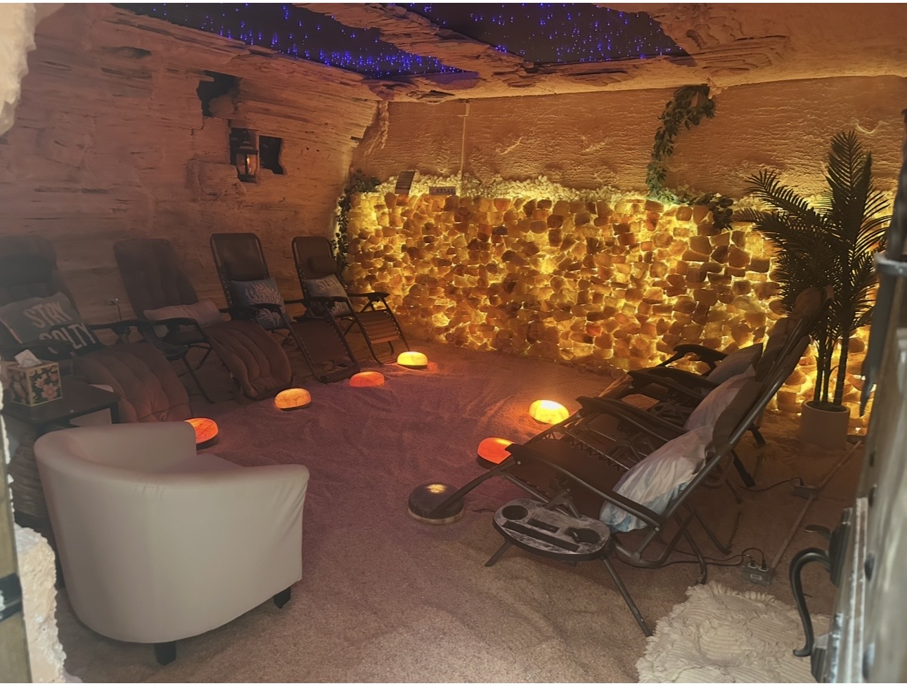 Indoor lounge with chairs, a white armchair, and a stone wall illuminated with warm lighting, plants, and a textured ceiling.