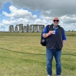 Person standing outdoors on grass, holding a drink, with Stonehenge in the background under cloudy sky.