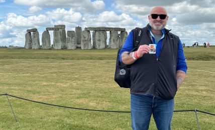 Person standing outdoors on grass, holding a drink, with Stonehenge in the background under cloudy sky.
