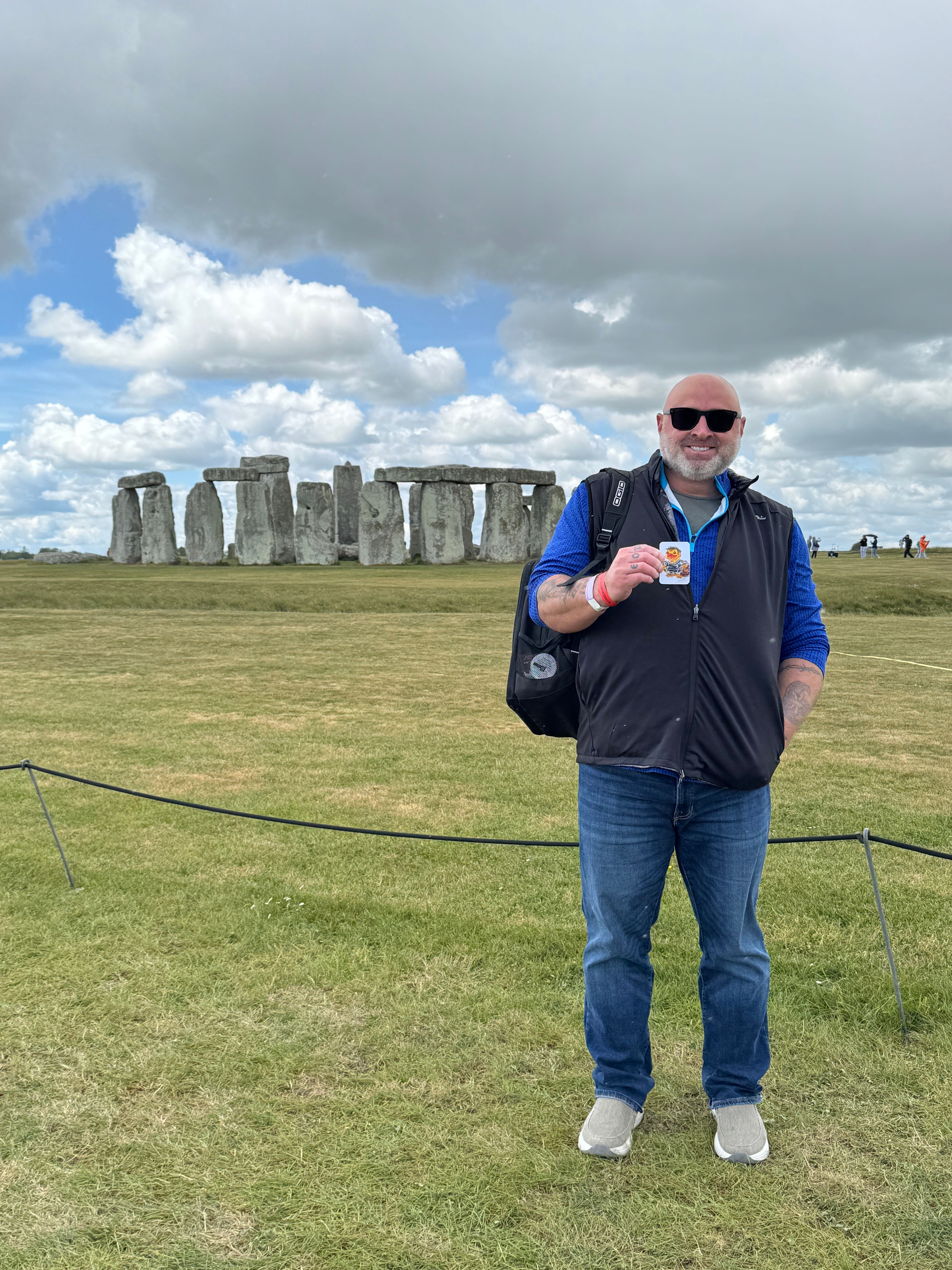 Person standing outdoors on grass, holding a drink, with Stonehenge in the background under cloudy sky.