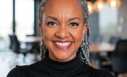 Smiling woman with gray hair styled in braids, wearing a black turtleneck, in a modern office setting with large windows and hanging lights.
