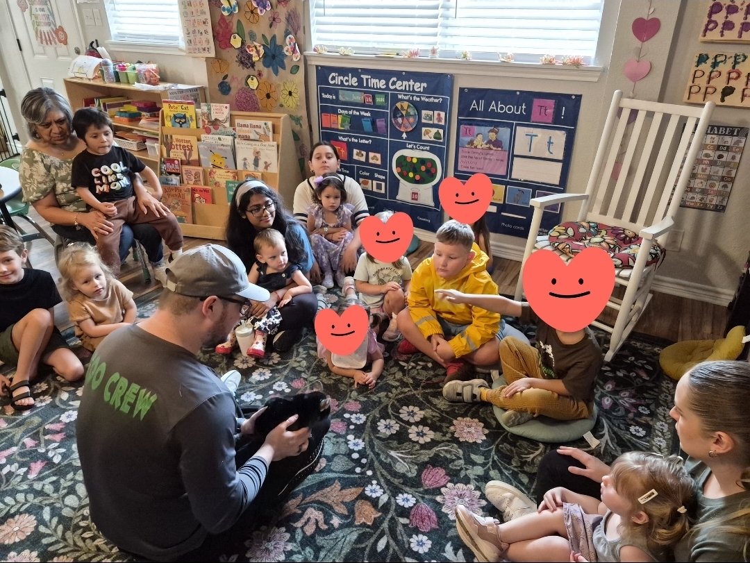 Group of children sitting on a colorful rug in a classroom, listening to an adult reading a book, with educational posters and toys around.
