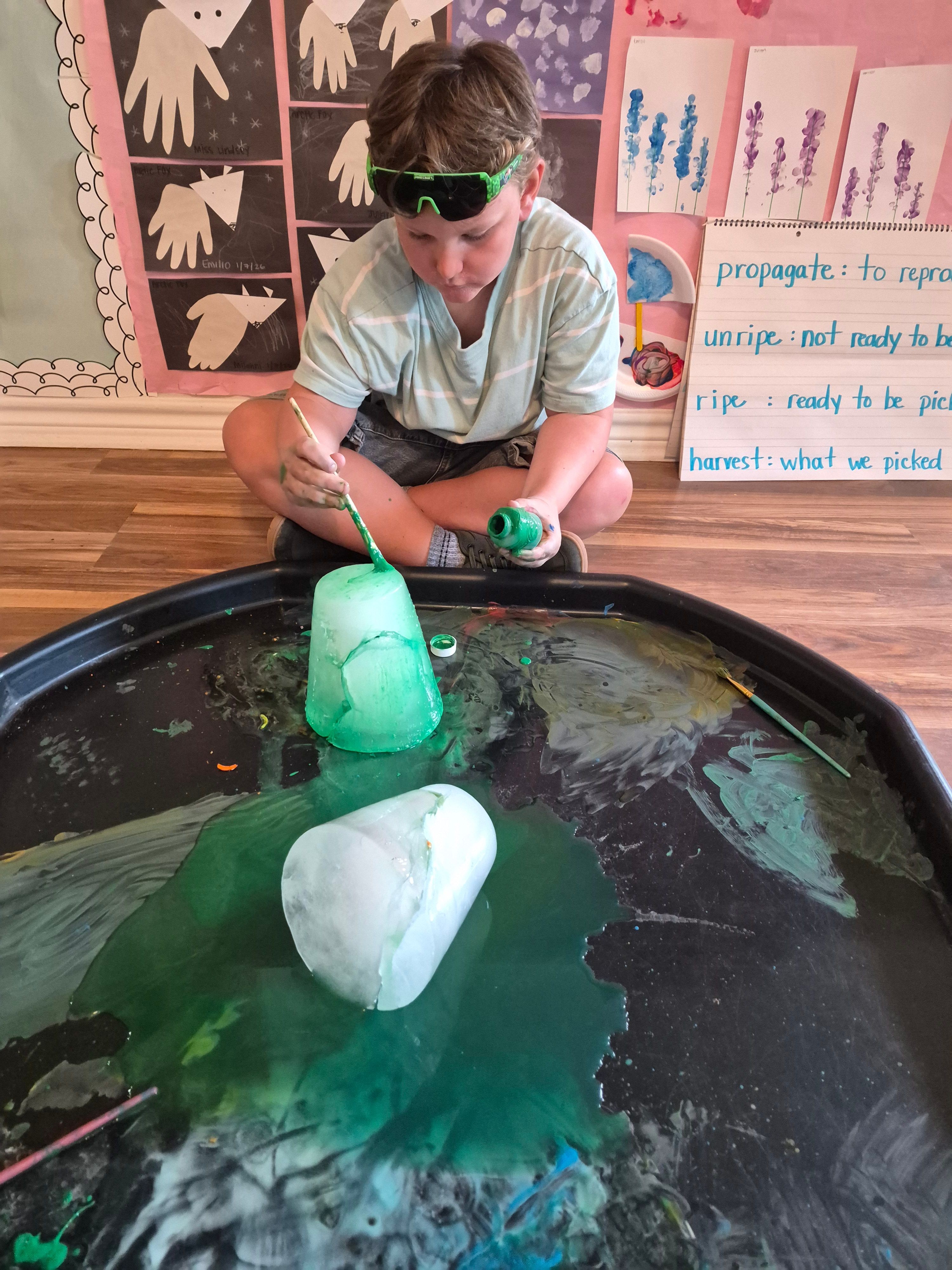 Child wearing safety goggles conducts a science experiment with balloons and liquids at a classroom table.