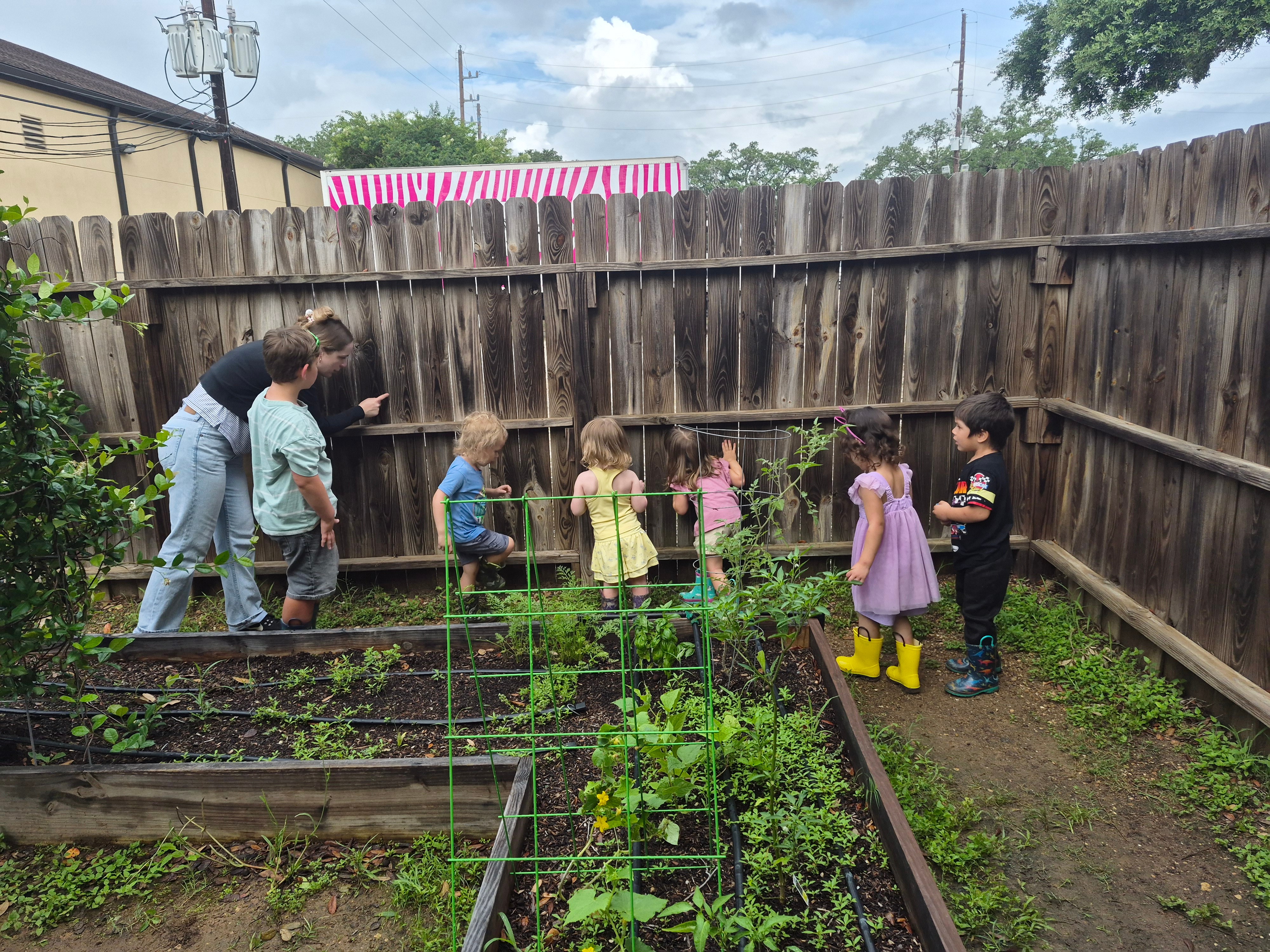 Group of children and adults in a garden with raised beds, some children wearing rain boots, some adults supervising.
