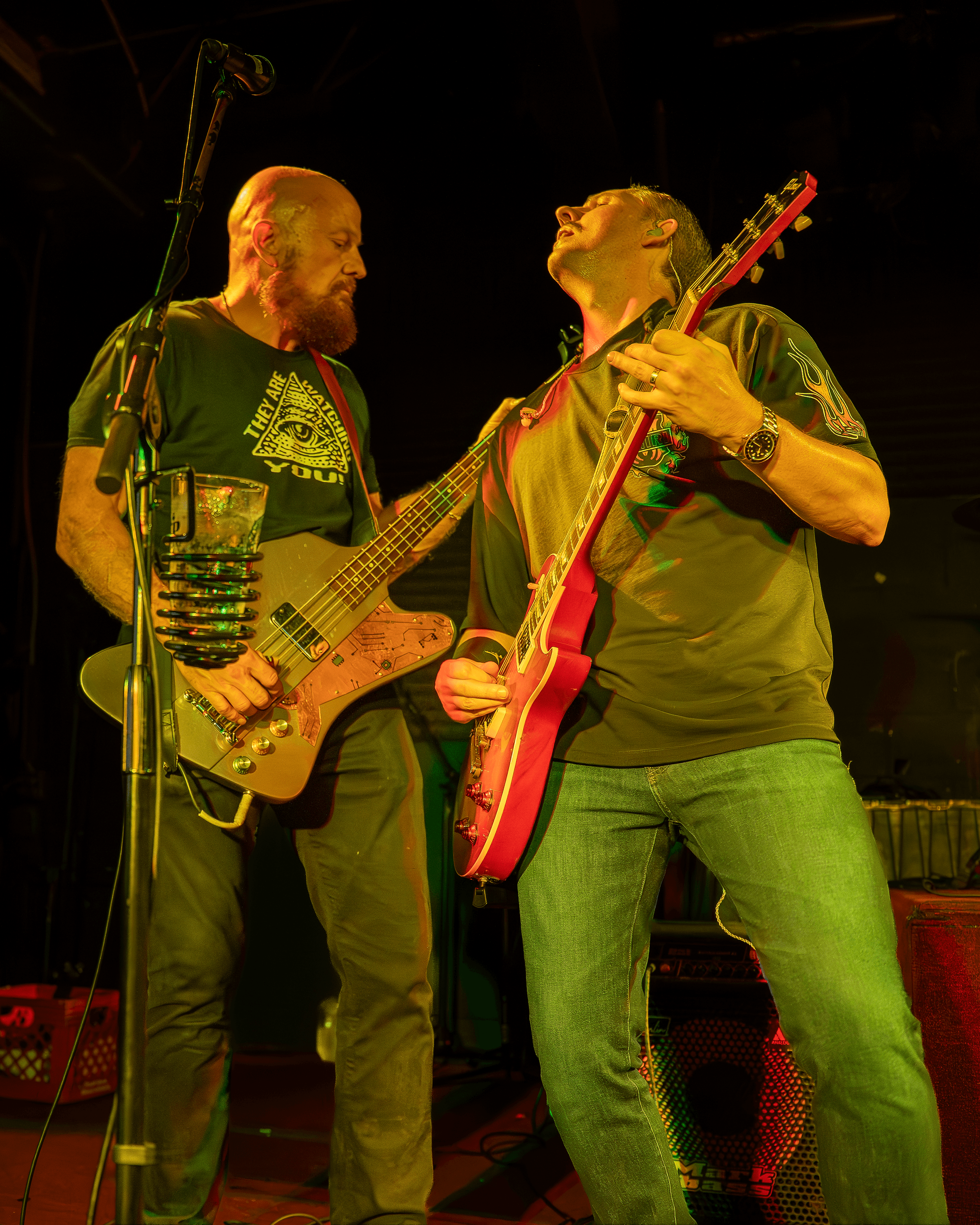 Two musicians playing guitars on stage, one with a glass of water, both engaged in performance, dark background.