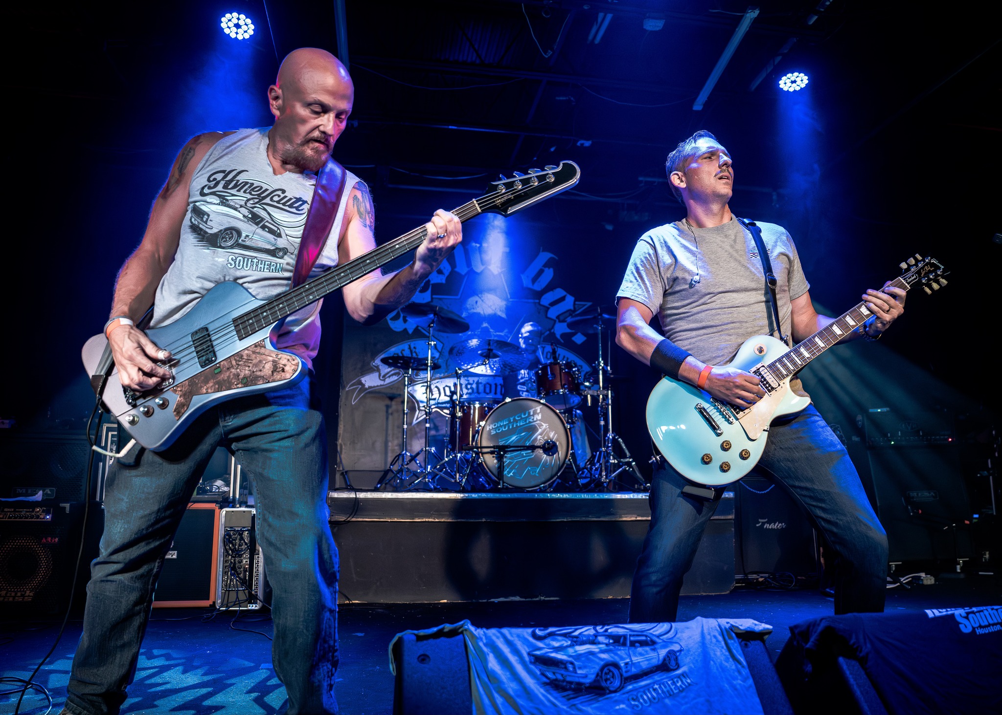 Two musicians playing electric guitars on stage with drums in the background, blue stage lighting, and a dark backdrop.