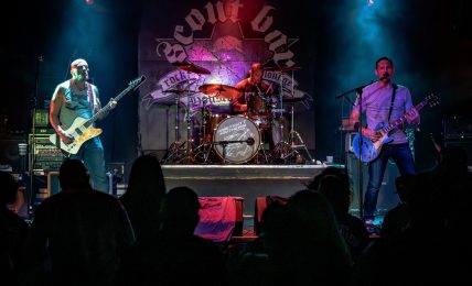 Band performs on stage with three musicians playing guitar and drums, audience silhouettes in foreground, stage backdrop with logo.