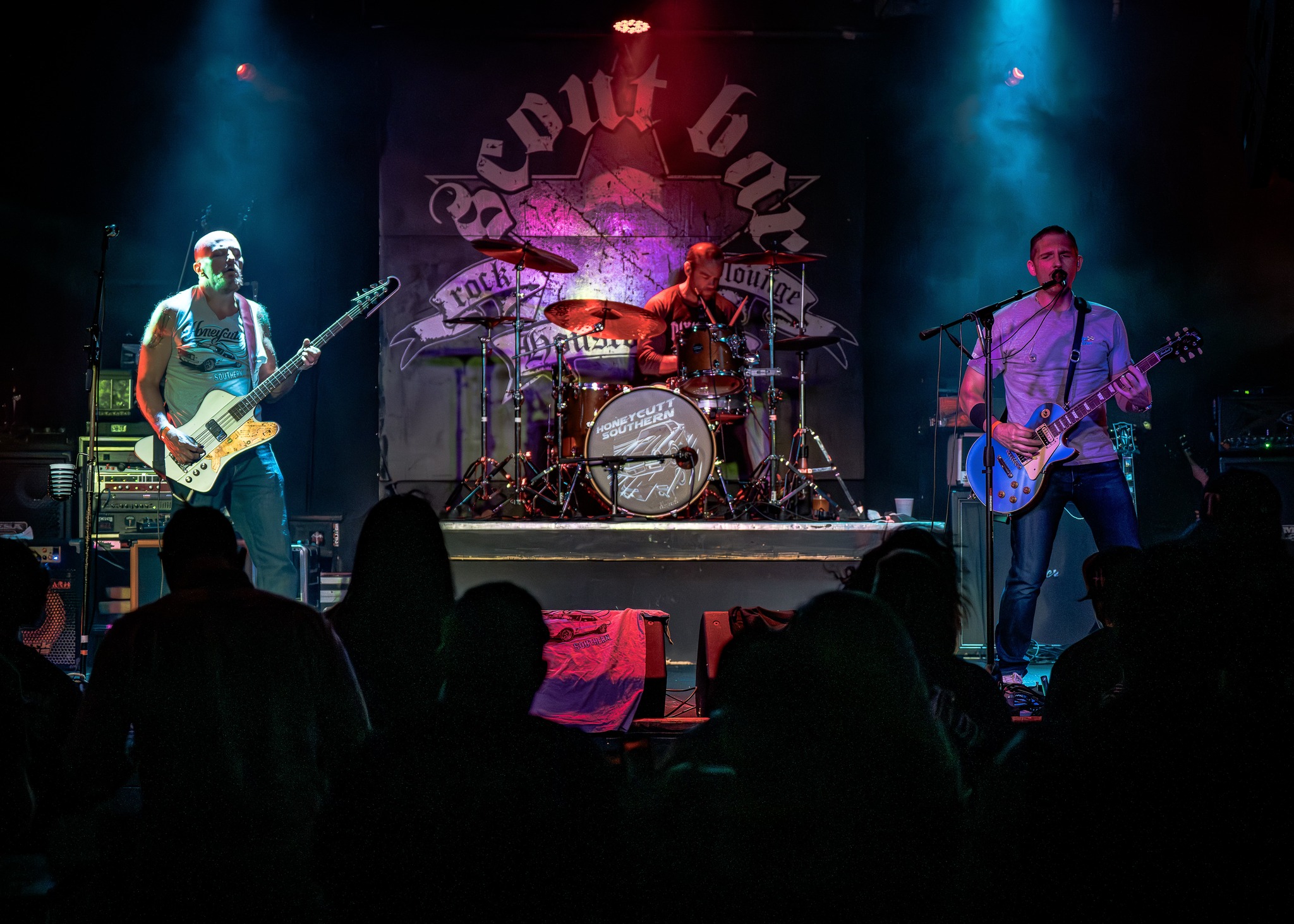 Band performs on stage with three musicians playing guitar and drums, audience silhouettes in foreground, stage backdrop with logo.