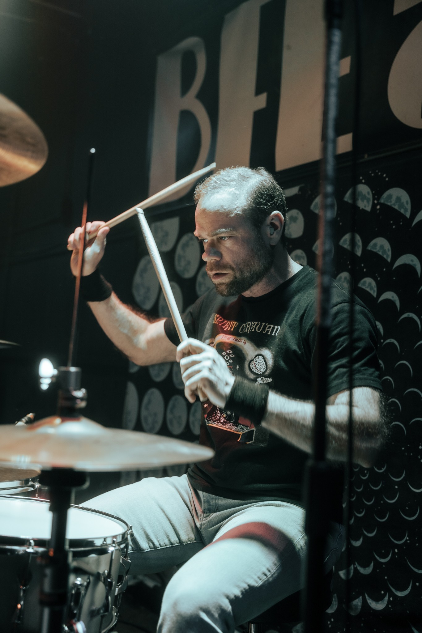 A man playing drums, holding drumsticks, with a focused expression, dark background with patterns, and large letters partially visible.