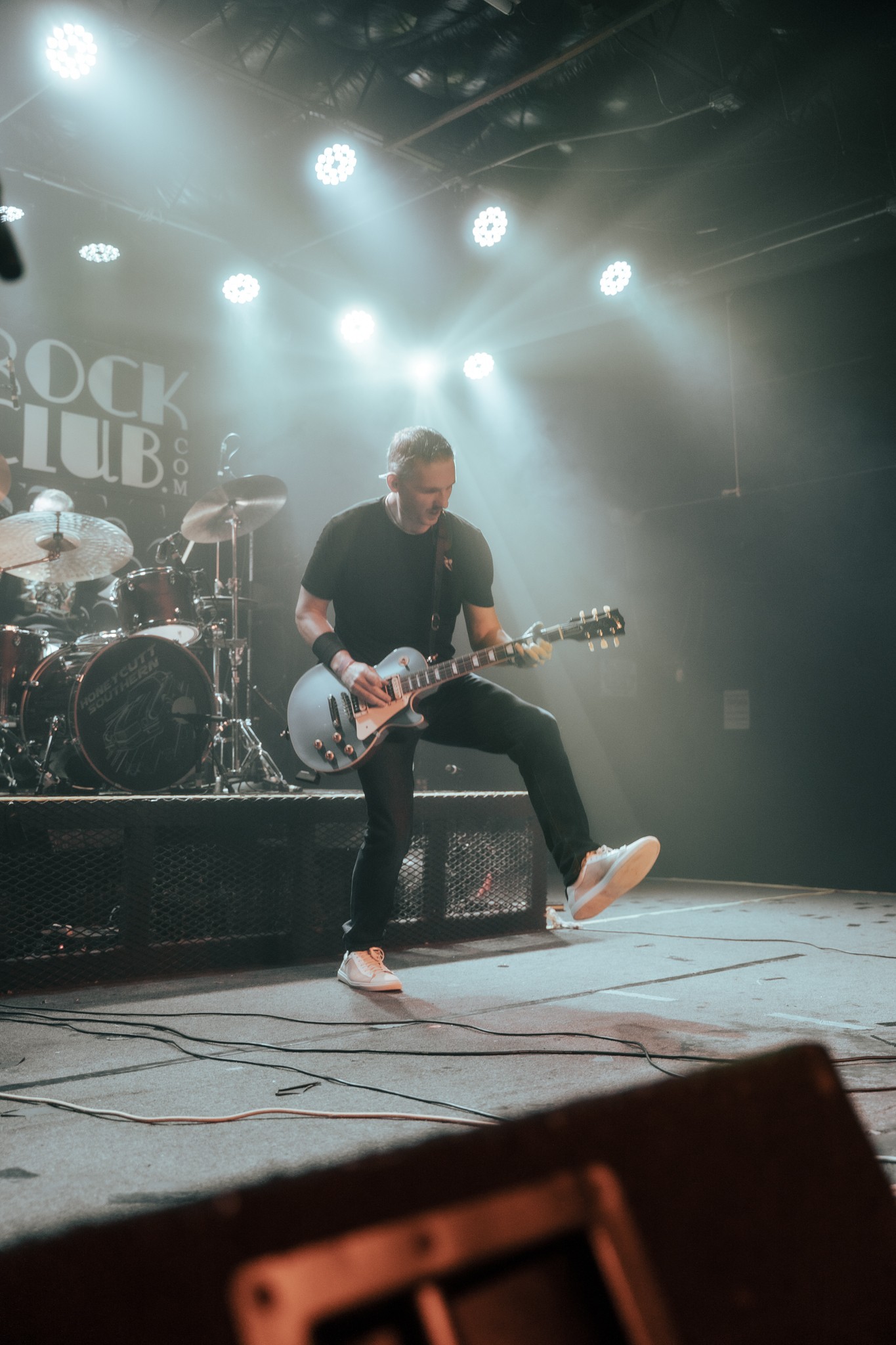 Guitarist performing on stage with bright lights, a drum set in the background, and a sign reading 'ROCK CLUB'.