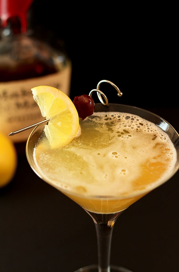 Cocktail glass with a lemon wedge and cherry garnish, filled with a frothy yellow drink, against a dark background.