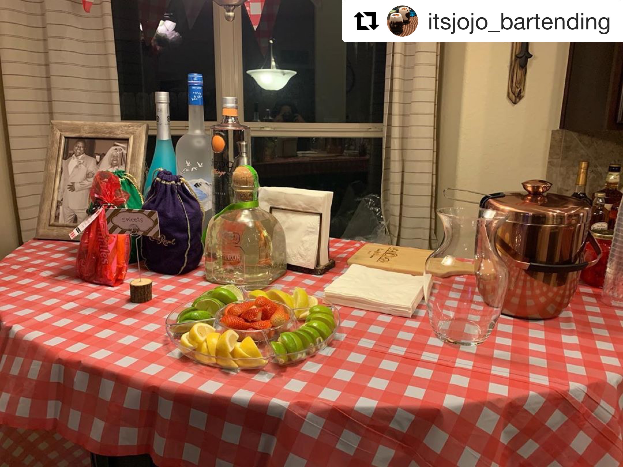 Table with red and white checkered tablecloth, various bottles, a fruit platter, and a copper container in a kitchen.