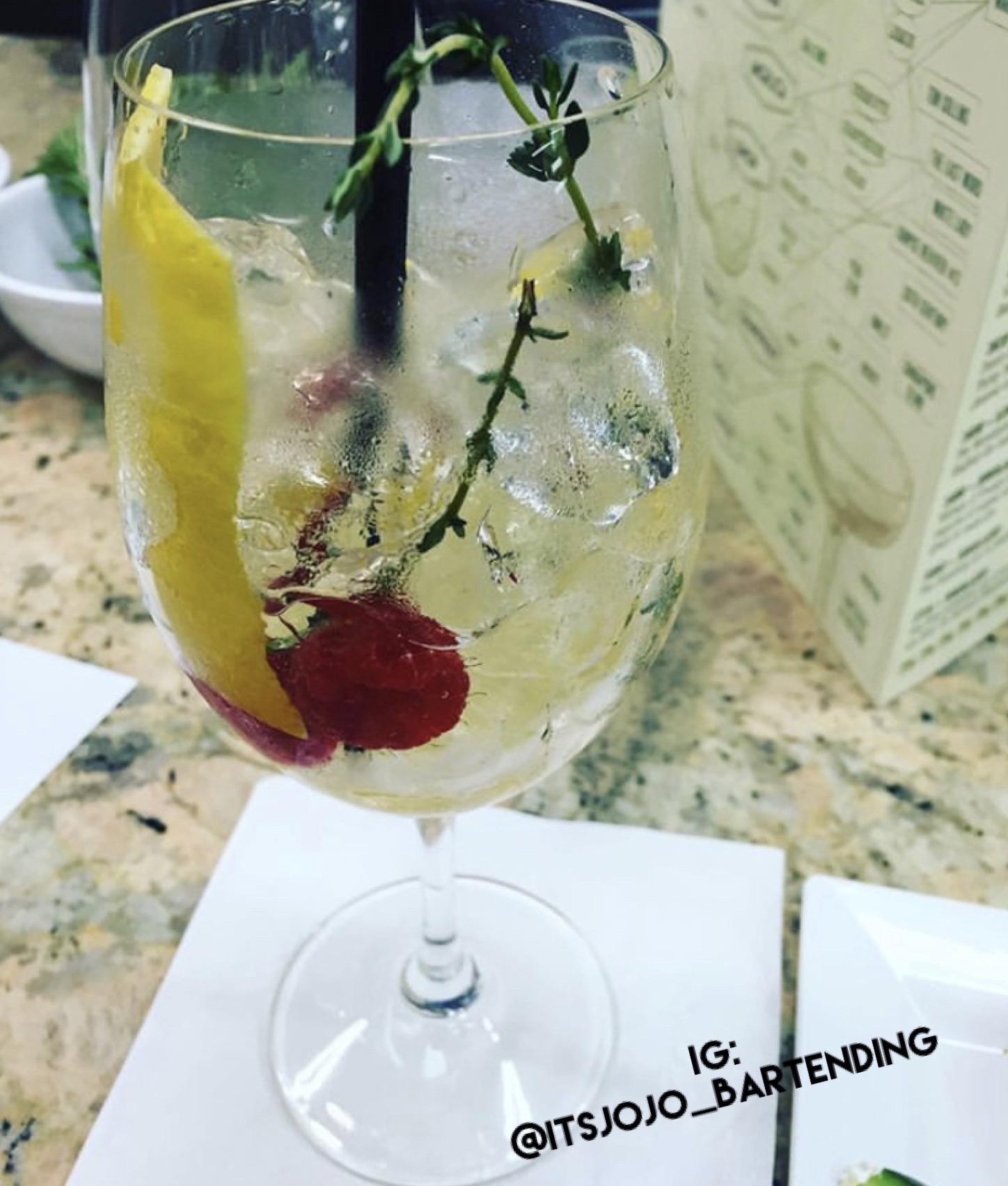 A glass with a lemon slice, cherry, ice, and herbs on a table, with a menu in the background.
