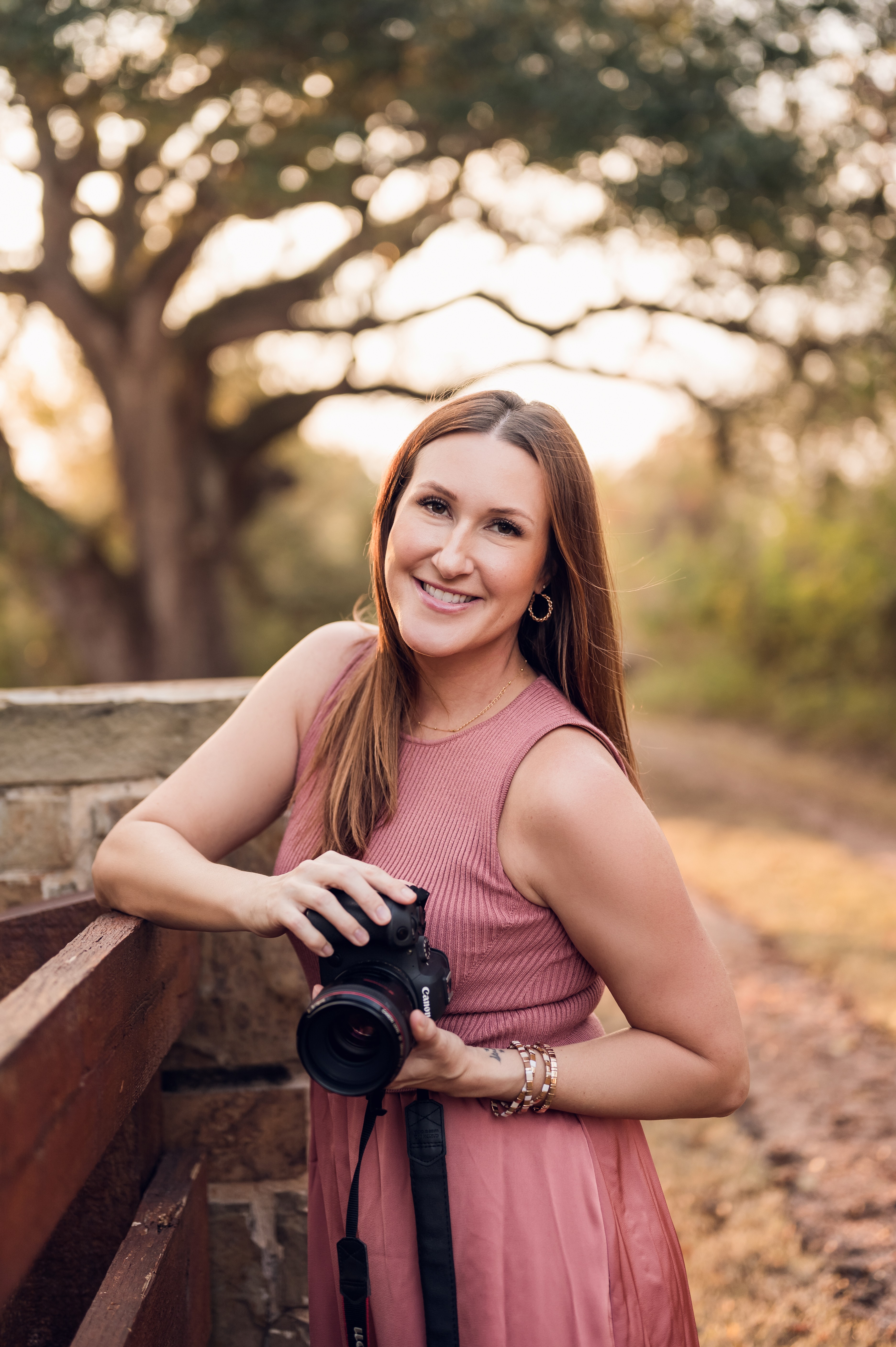 Smiling woman holding a camera outdoors in a park with trees and a dirt path, wearing a pink dress.