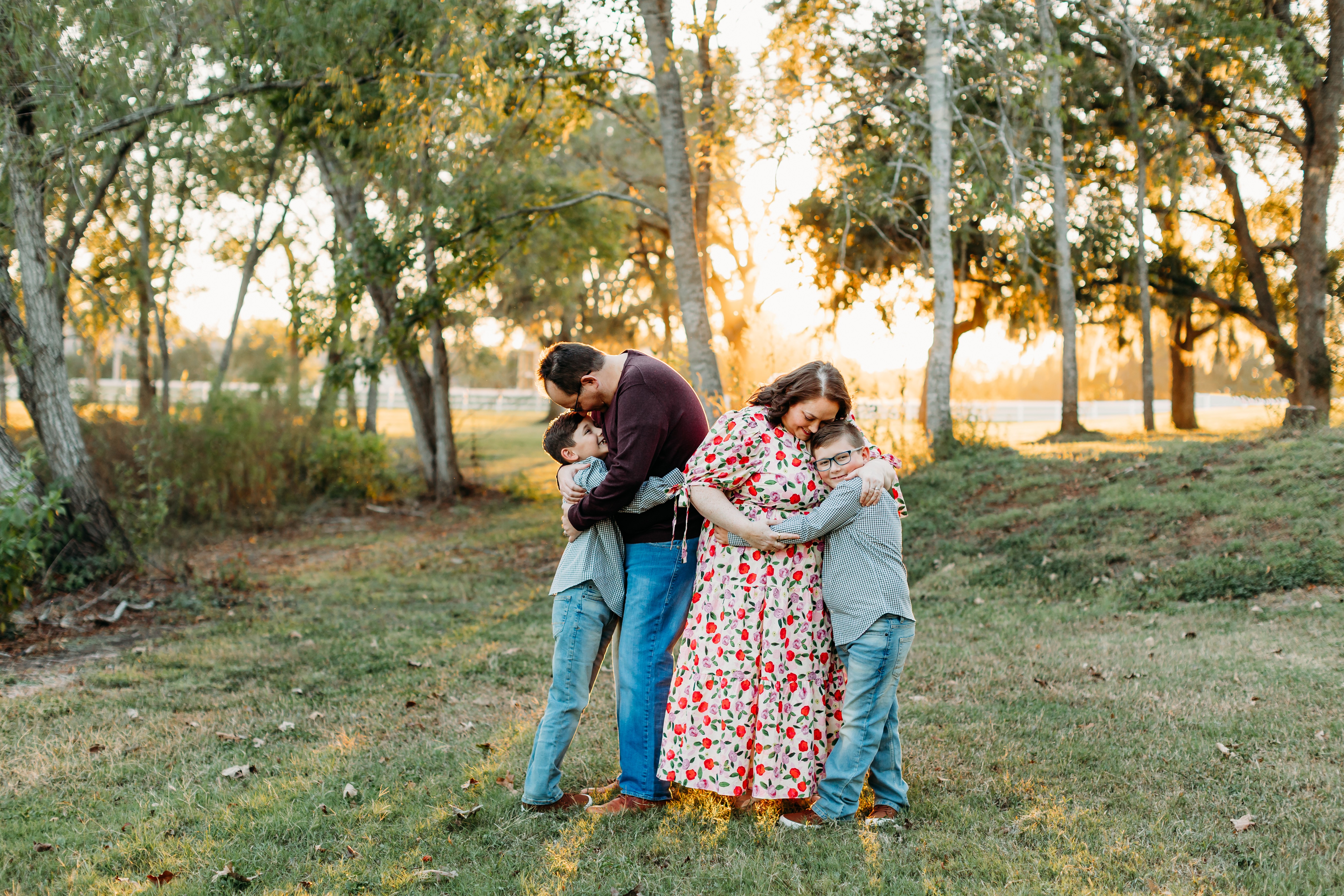 Two women and a child hugging outdoors in a park during sunset, trees in the background.