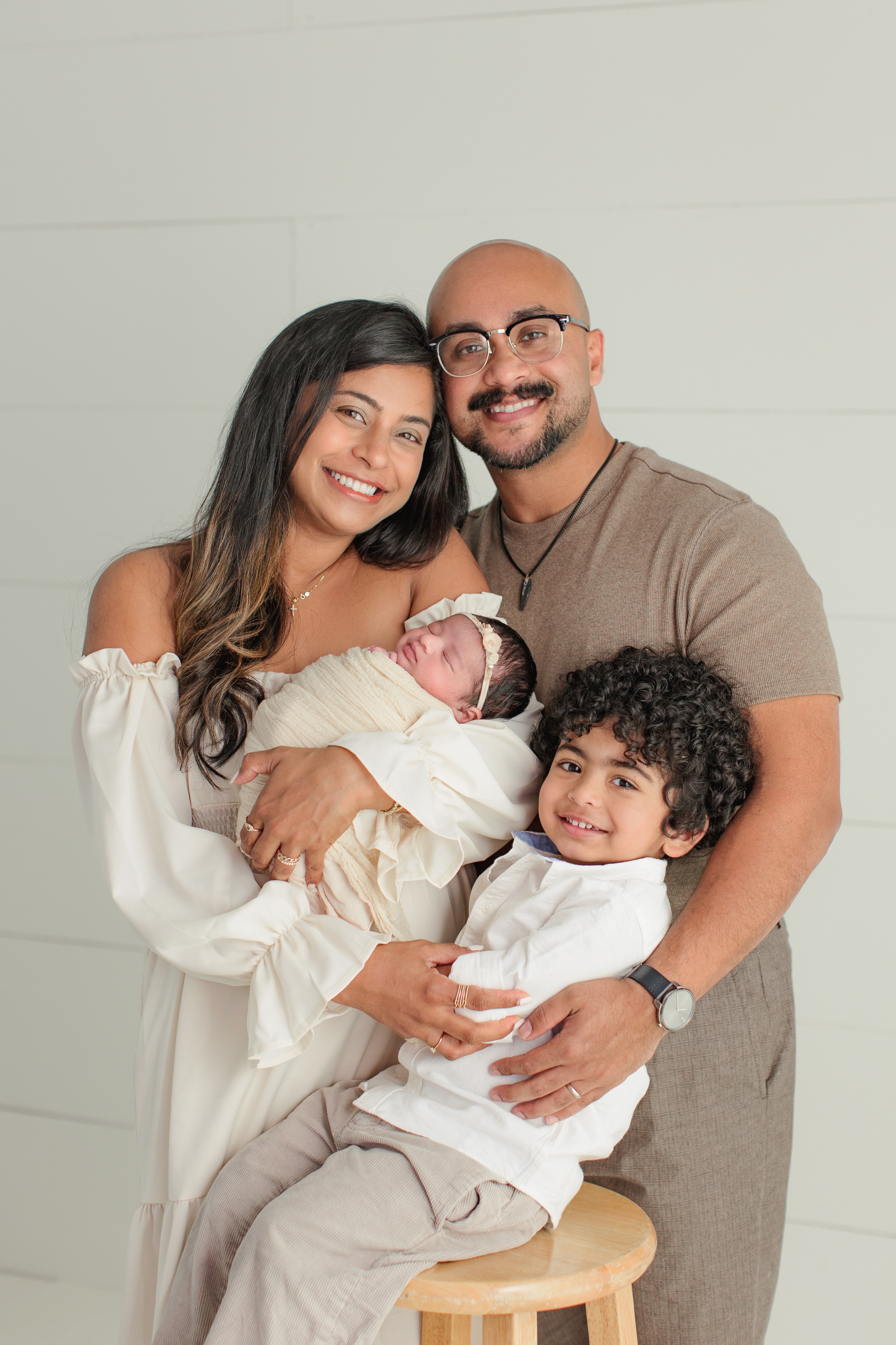 Family of four smiling, with a woman holding a newborn, a man, and a young girl, against a plain background.