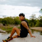 Person sitting on a rock near water with trees and cloudy sky in background, wearing black clothing and colorful shoes.