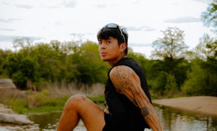 Person sitting on a rock near water with trees and cloudy sky in background, wearing black clothing and colorful shoes.