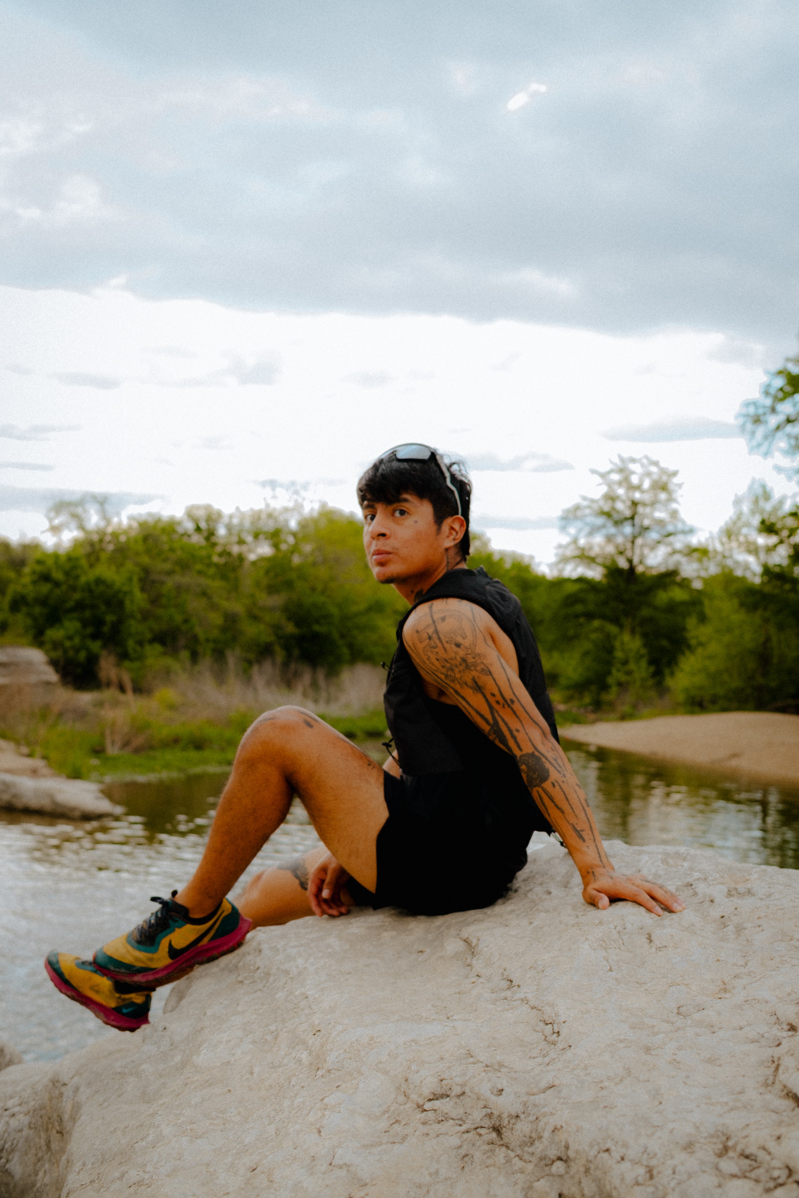 Person sitting on a rock near water with trees and cloudy sky in background, wearing black clothing and colorful shoes.