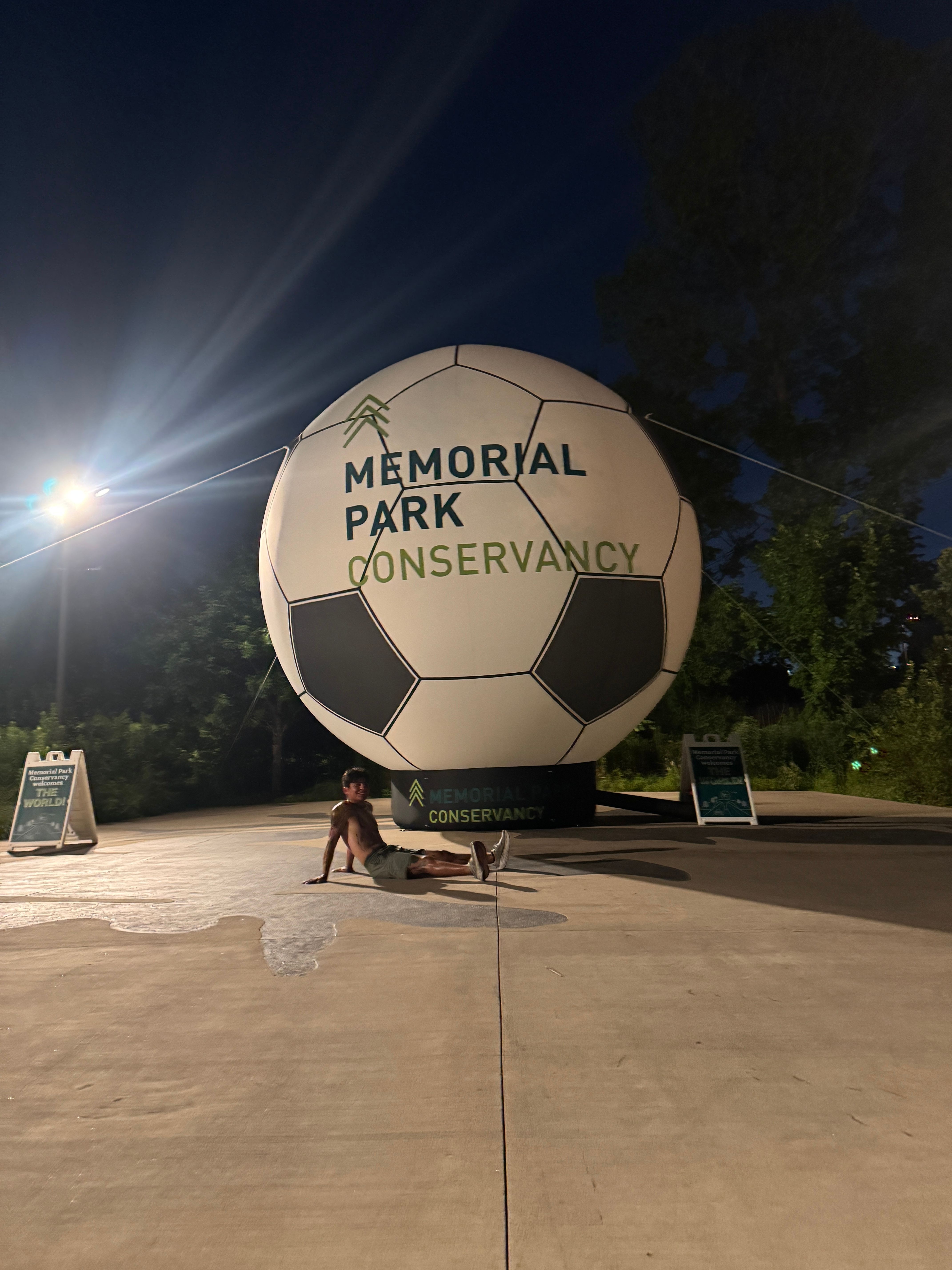 Person sitting on the ground near a large soccer ball-shaped memorial with text, on a concrete surface, trees in background.