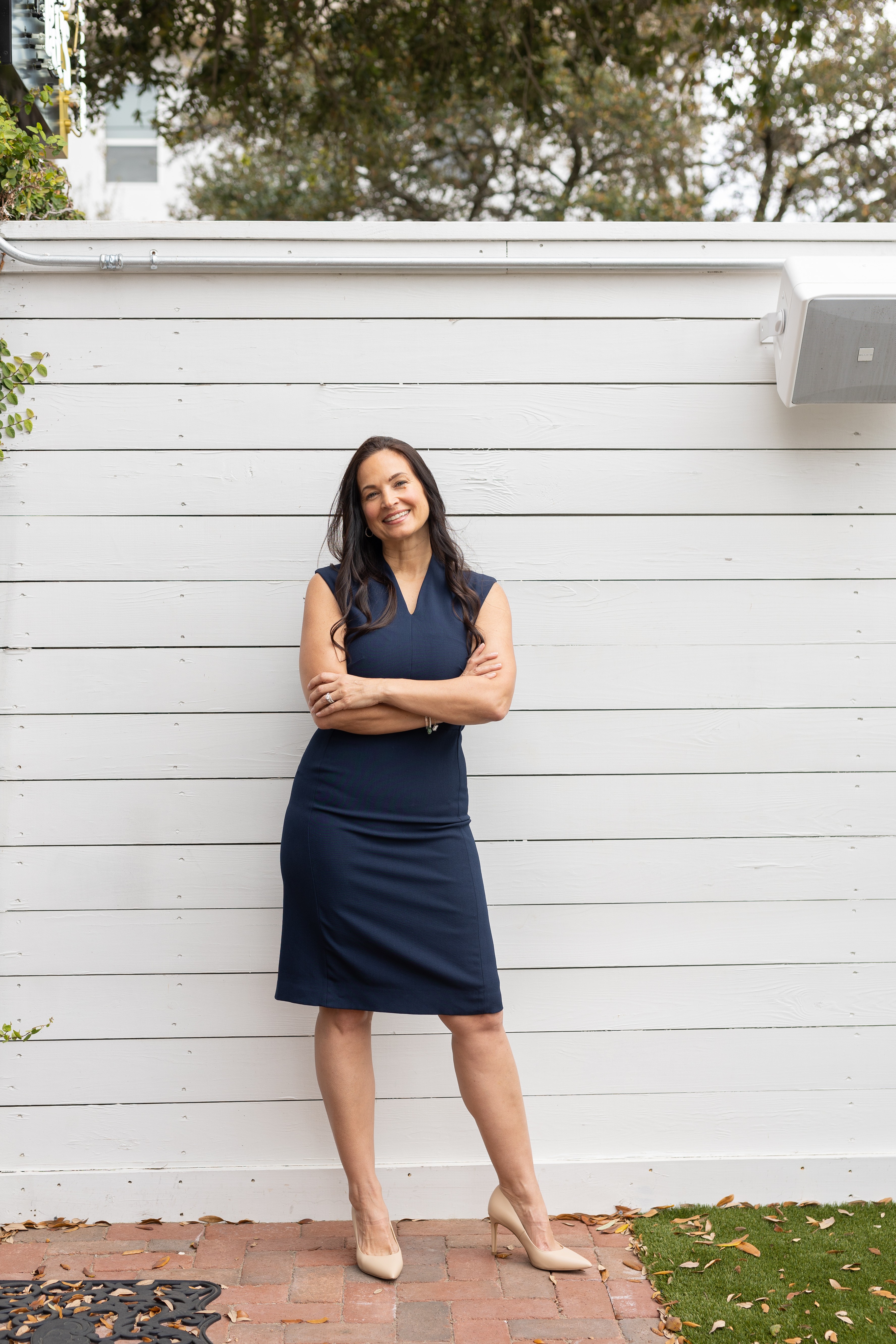 Woman standing with arms crossed in front of a white wooden fence outdoors, smiling, wearing a navy dress and heels.