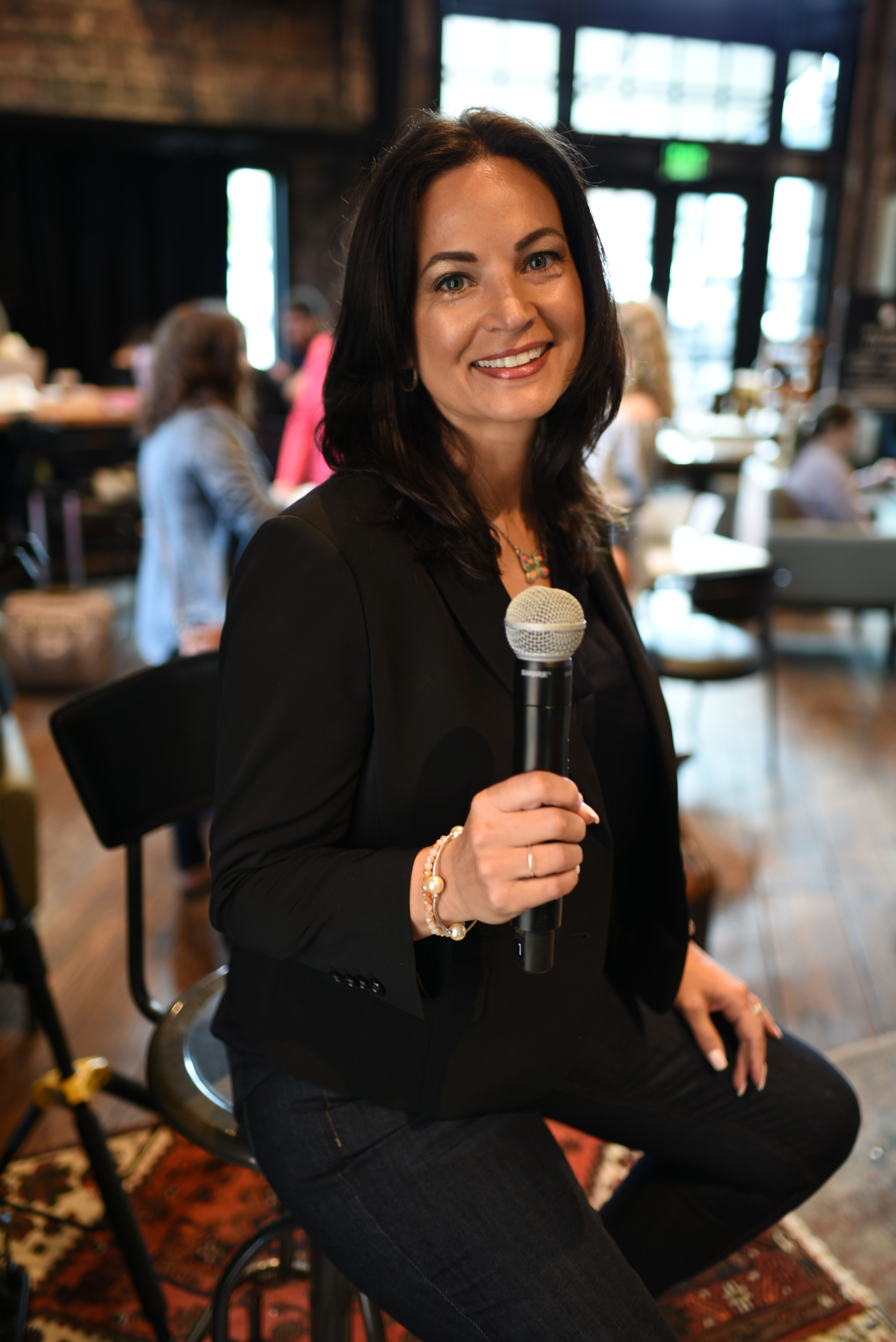 Woman with long dark hair smiling, holding a microphone, wearing a black blazer, in a lively indoor setting.