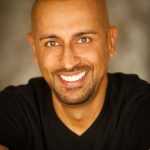 Man with a shaved head and beard smiling, wearing a black shirt and bracelets, against a blurred background.