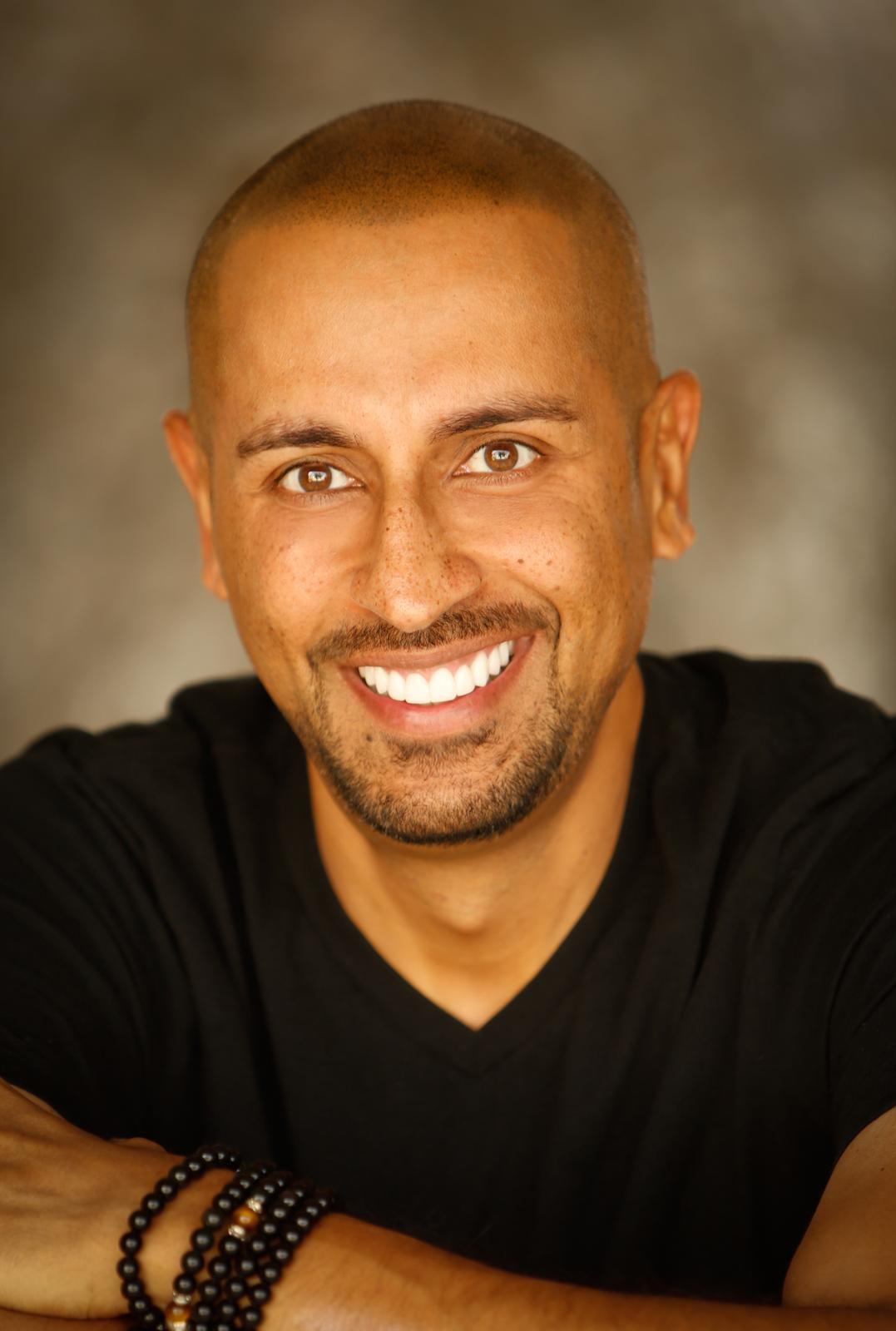 Man with a shaved head and beard smiling, wearing a black shirt and bracelets, against a blurred background.