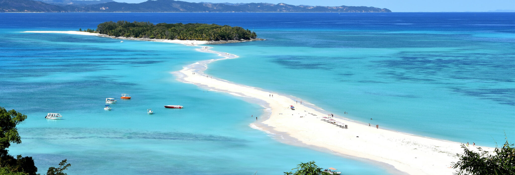 A tropical beach with white sand, turquoise water, boats, and an island in the distance.