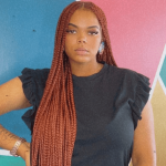 Young woman with long braided hair wearing a black top with ruffled sleeves, standing against a colorful geometric background.