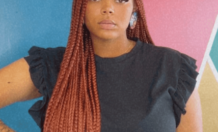 Young woman with long braided hair wearing a black top with ruffled sleeves, standing against a colorful geometric background.