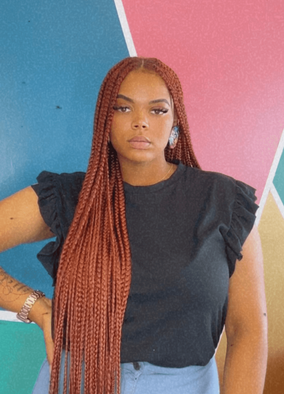 Young woman with long braided hair wearing a black top with ruffled sleeves, standing against a colorful geometric background.