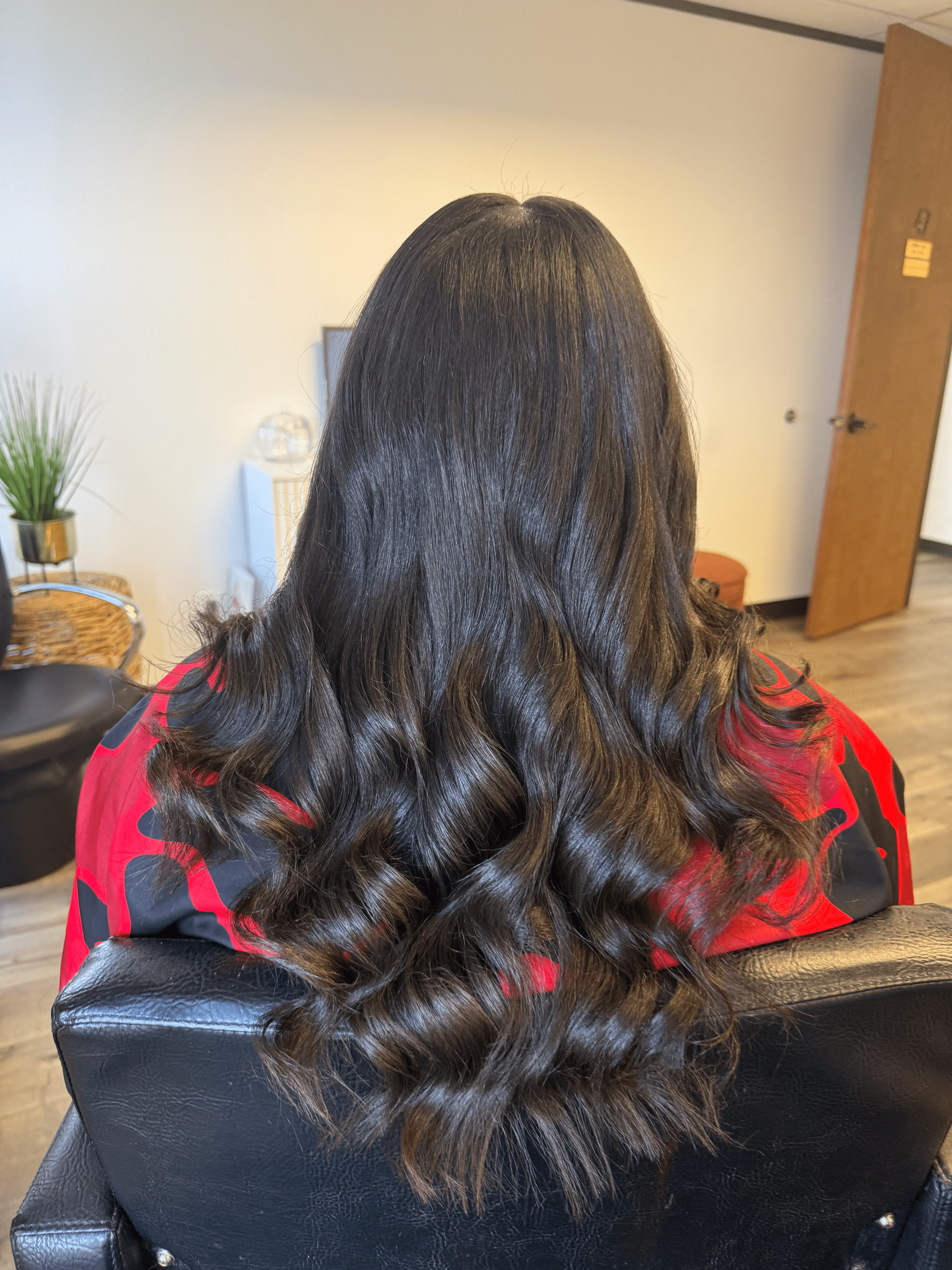Back view of a person with long, dark, wavy hair wearing a red and black checkered shirt, sitting in a room.