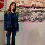 Young woman with long hair in a blue jacket and jeans standing in front of abstract artwork on wall.