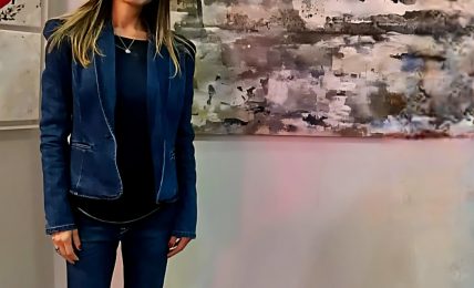 Young woman with long hair in a blue jacket and jeans standing in front of abstract artwork on wall.