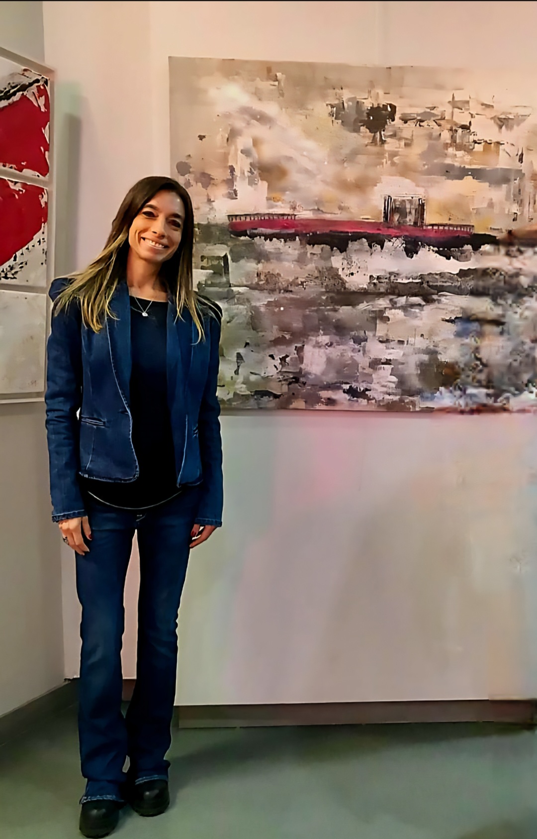 Young woman with long hair in a blue jacket and jeans standing in front of abstract artwork on wall.