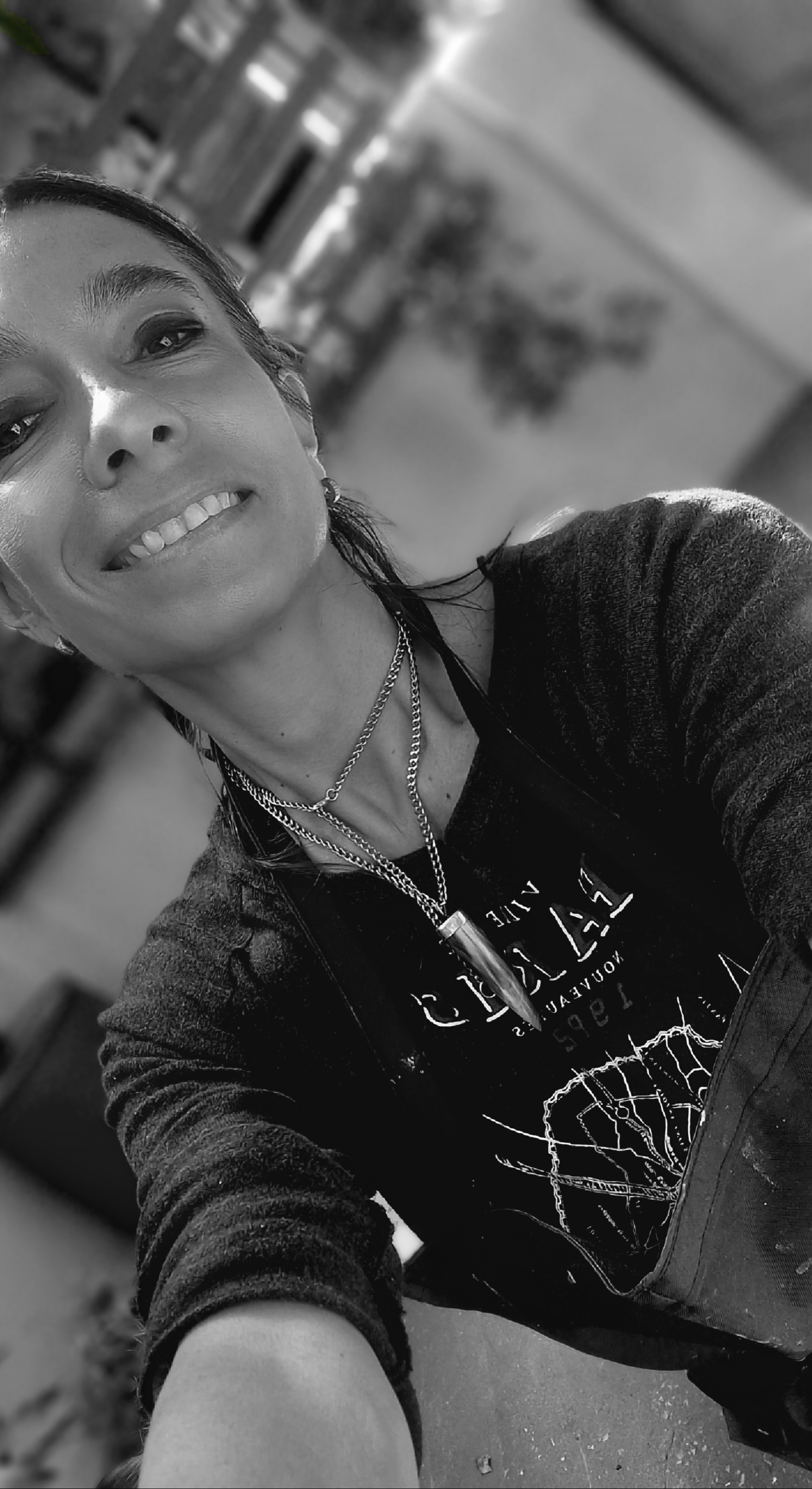 Person smiling, wearing a dark top and layered necklaces, in an indoor setting with blurred background.