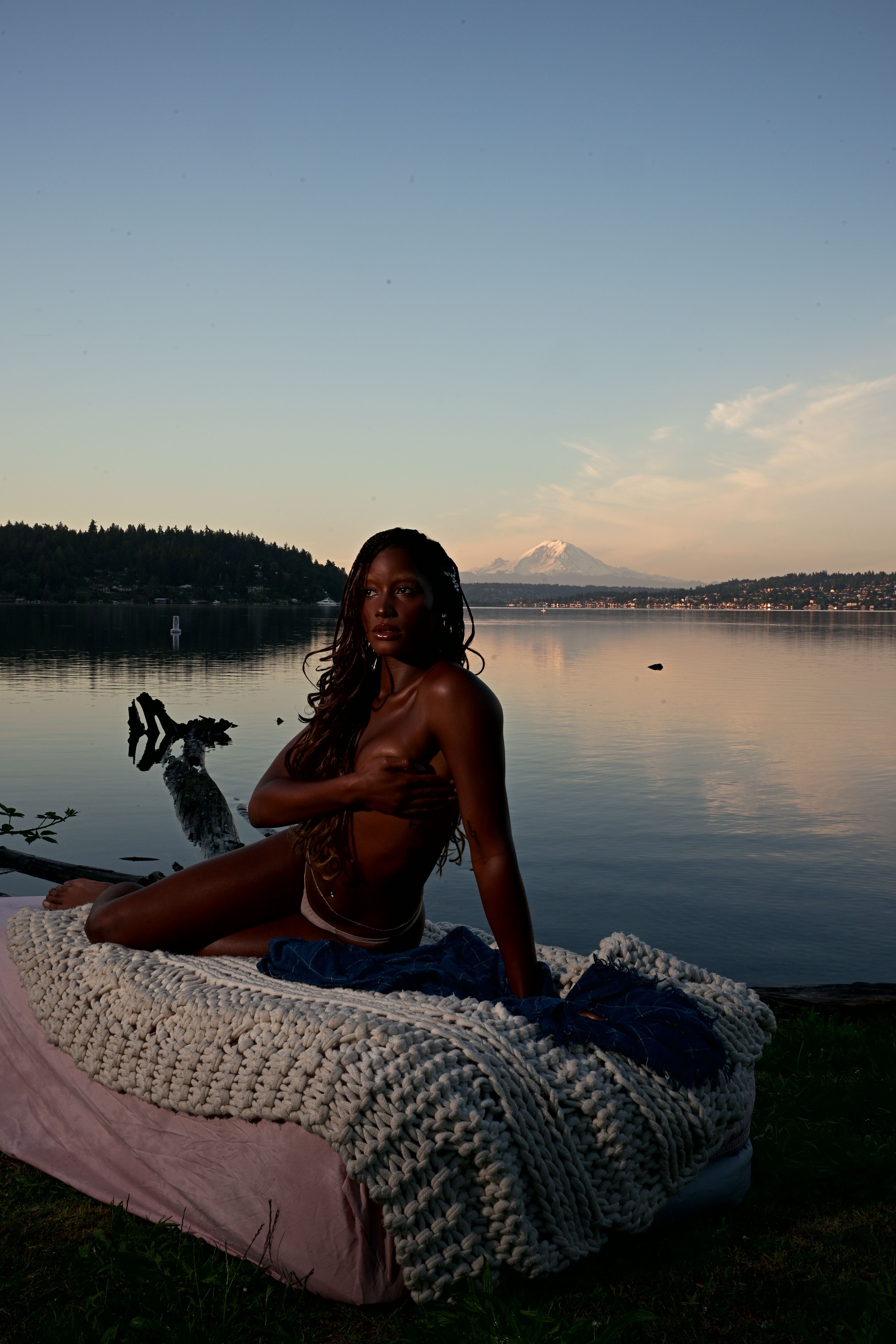 Person sitting on a blanket by the water during sunset, with a bicycle in the background, and a cloudy sky.