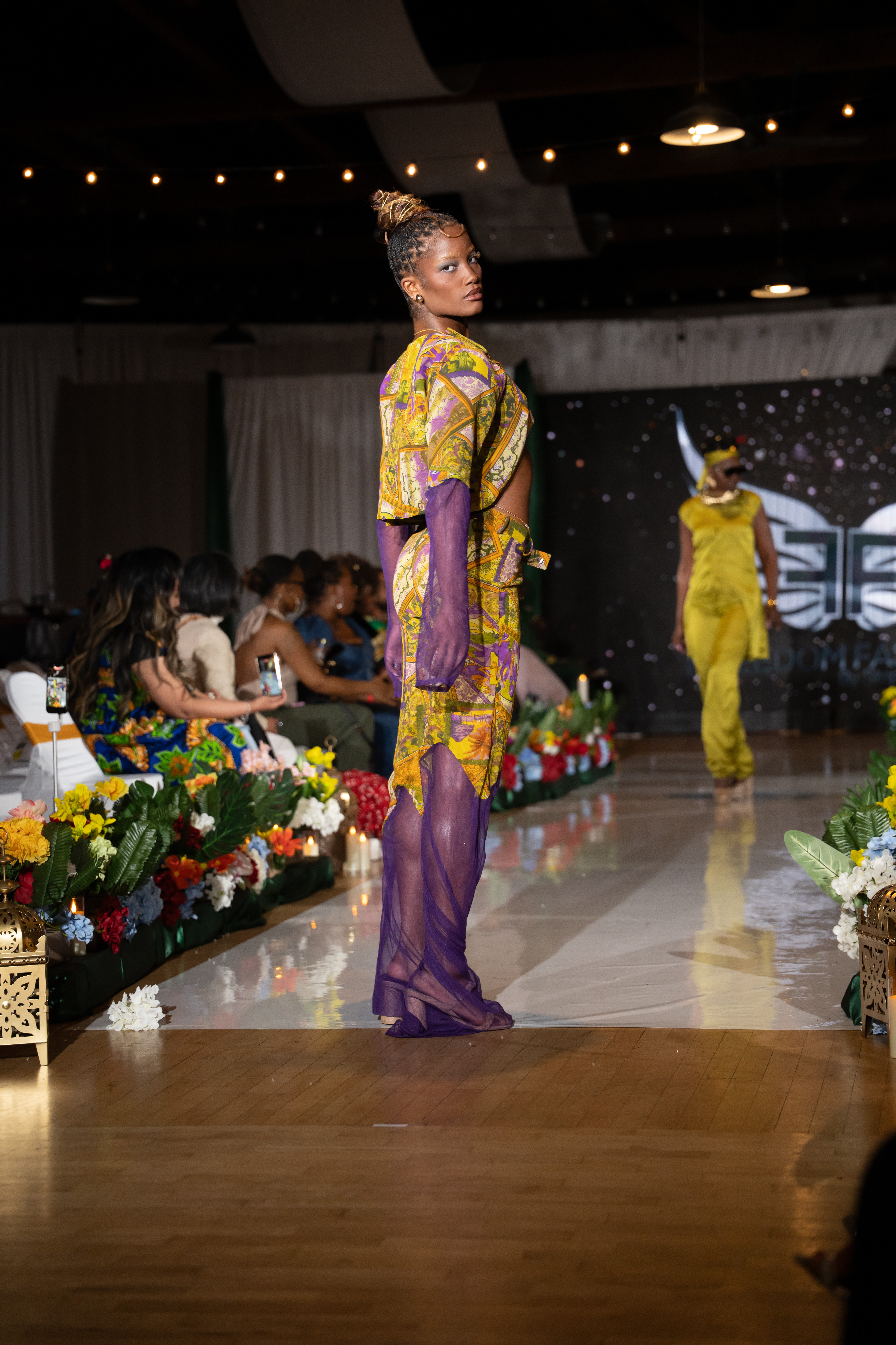 Model in colorful traditional outfit walking on runway with audience and floral decorations, stage background visible.