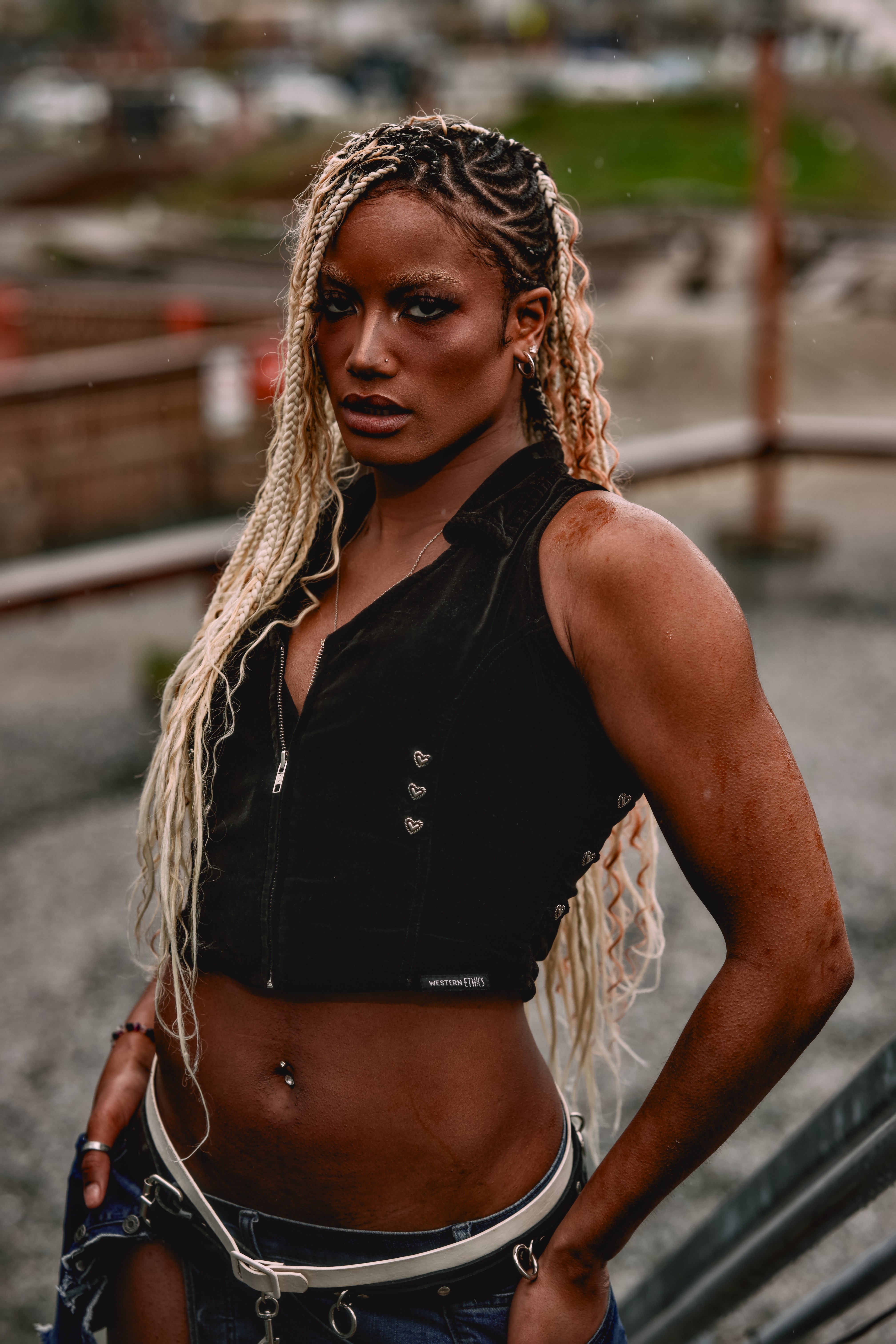 Woman with long braided hair and dark makeup, wearing a sleeveless black top, standing outdoors with a blurred background.