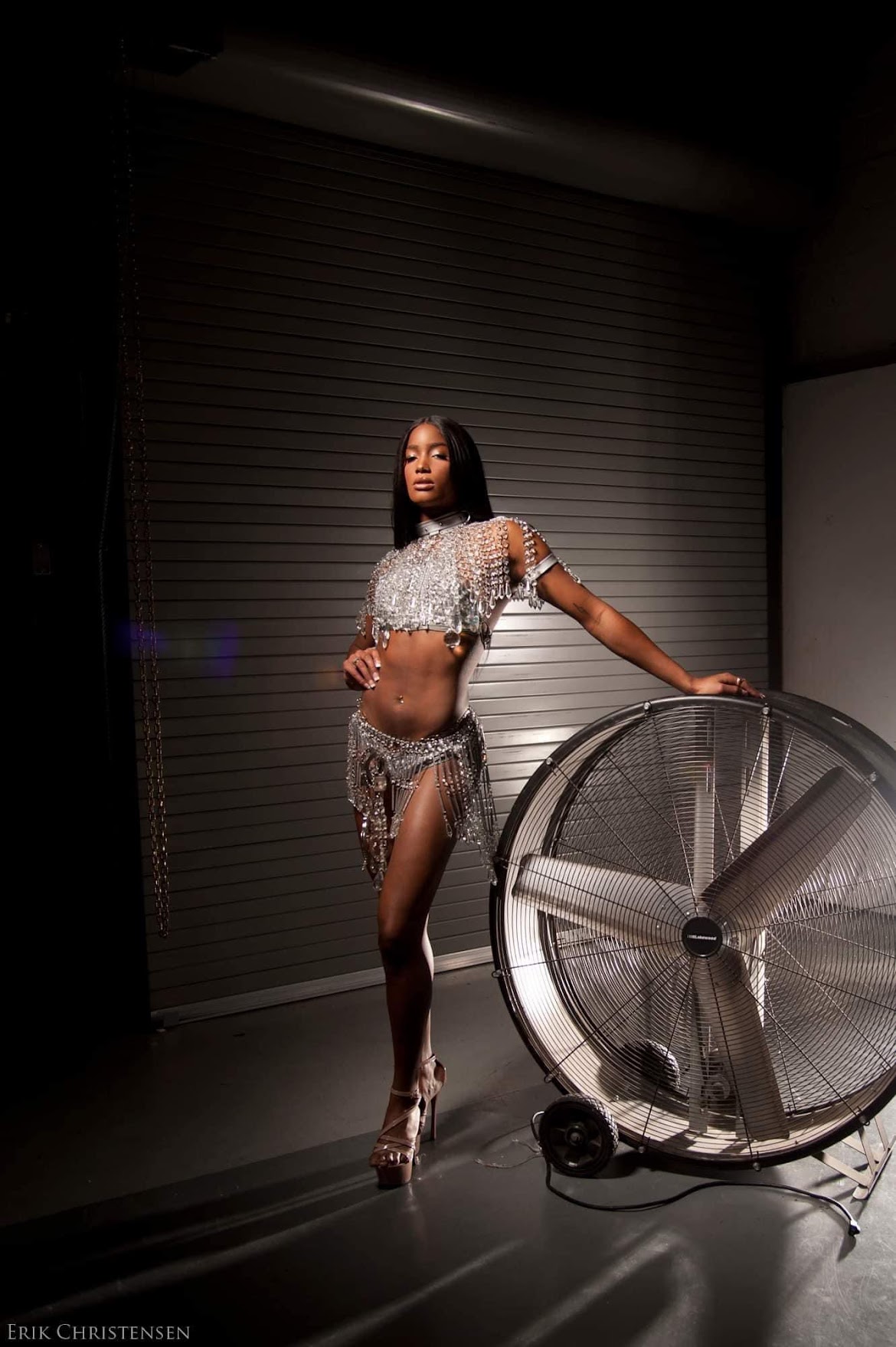 A woman in a patterned outfit poses next to a large industrial fan in a dimly lit room.