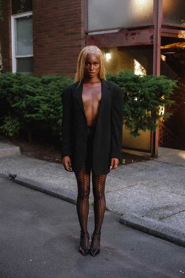Woman standing outdoors in a black blazer, fishnet stockings, and high heels, with a building and greenery behind her.