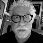 Portrait of an older man with curly white hair, beard, and glasses, in a room with framed pictures on the wall.